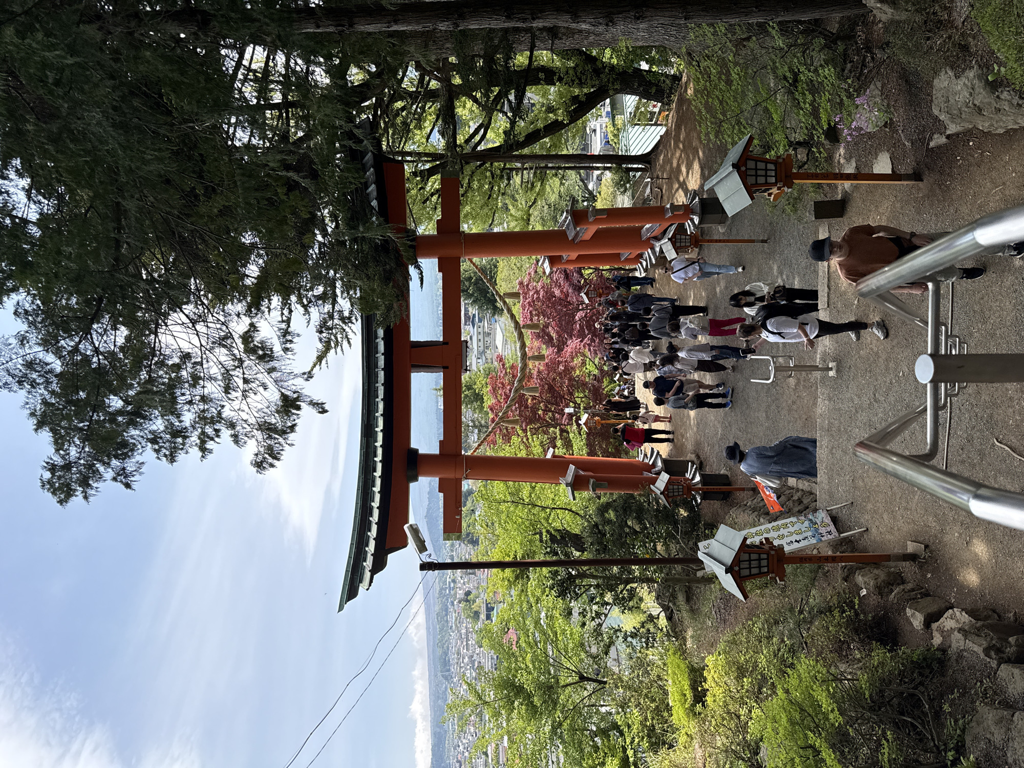 A vibrant outdoor scene depicts the entrance to a Japanese shrine, identified by the visible text and the distinct torii gate, located in Fujiyoshida, Japan. The image, oriented vertically, shows a wide view of a paved or dirt path leading up to a large, vermilion-colored torii gate, with a cityscape visible in the distant valley below.
Numerous people are gathered on the path, some standing in groups, others appearing to walk or queue. They are dressed in casual attire, suggesting visitors or tourists. To the right, a man in an orange shirt and black cap is visible near a metal railing, which likely borders a set of steps or a sloped ascent. Traditional wooden lanterns with small roofs and paper signs are placed along the path, guiding the way.
The surroundings are lush with green foliage and tall trees, some with reddish leaves, framing the scene. A rope, possibly a shimenawa, is strung across the top of the torii gate or nearby trees. The sky is clear and blue with sparse clouds, indicating good weather during the day. Parts of traditional Japanese architecture, including green-tiled roofs, are partially visible on the left. A utility pole and wires are also present.
Visible text from the scene reads: "神社の境内です。" (This is the shrine grounds.), "1リをしましょう" (which likely means "Let's pay a visit to the shrine."), and "富士表司神社" (Fujiyoshida Shrine), confirming the specific location. The scene captures a moment of cultural visitation or pilgrimage at a scenic Japanese shrine overlooking a town.