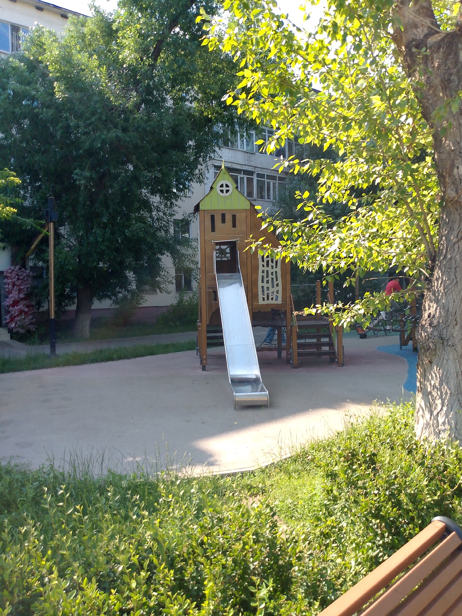 In Astana, Kazakhstan, a sunlit playground features a wooden play structure with a silver slide. The play structure is situated on a paved surface, surrounded by green grass and bushes. A large tree with sun-dappled leaves is prominent in the foreground. In the background, a multi-story building with windows is visible, partially obscured by foliage. A portion of a brown wooden bench is visible in the bottom right corner.