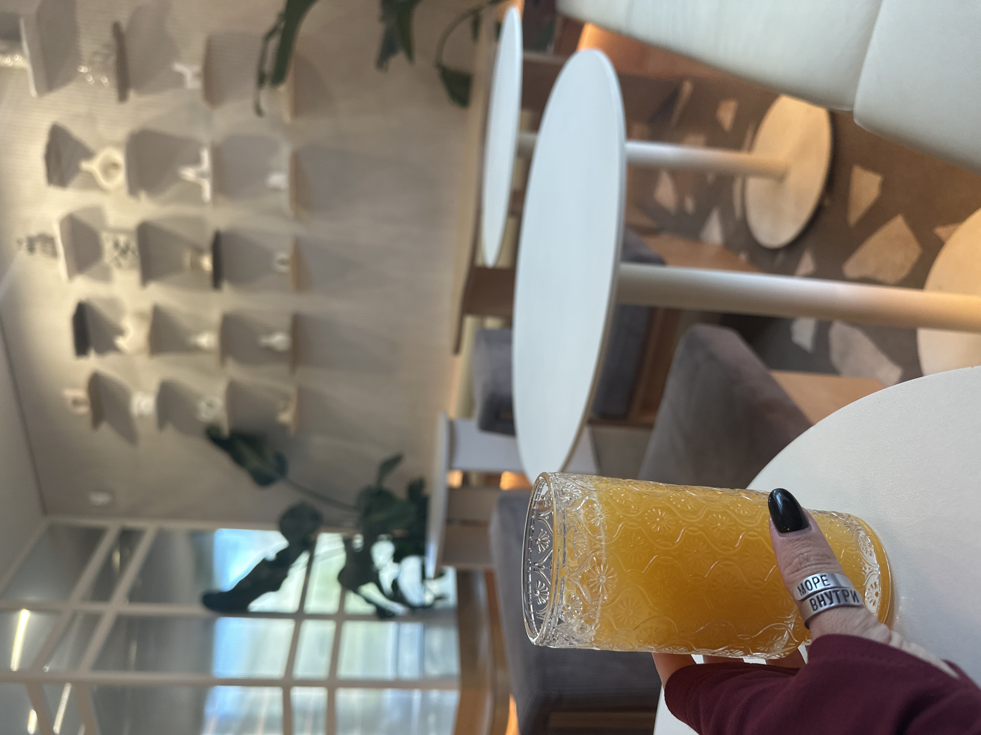 A person with a ring on their finger is holding a glass of orange juice in a cafe, near a white table. The cafe has an interesting architectural design with a wall decorated with multiple geometric shapes, resembling stylized shelves or display areas. In the background, there is a window with greenery visible, adding a touch of nature to the interior. The flooring is of light stone. This is Novosibirsk, Russia.