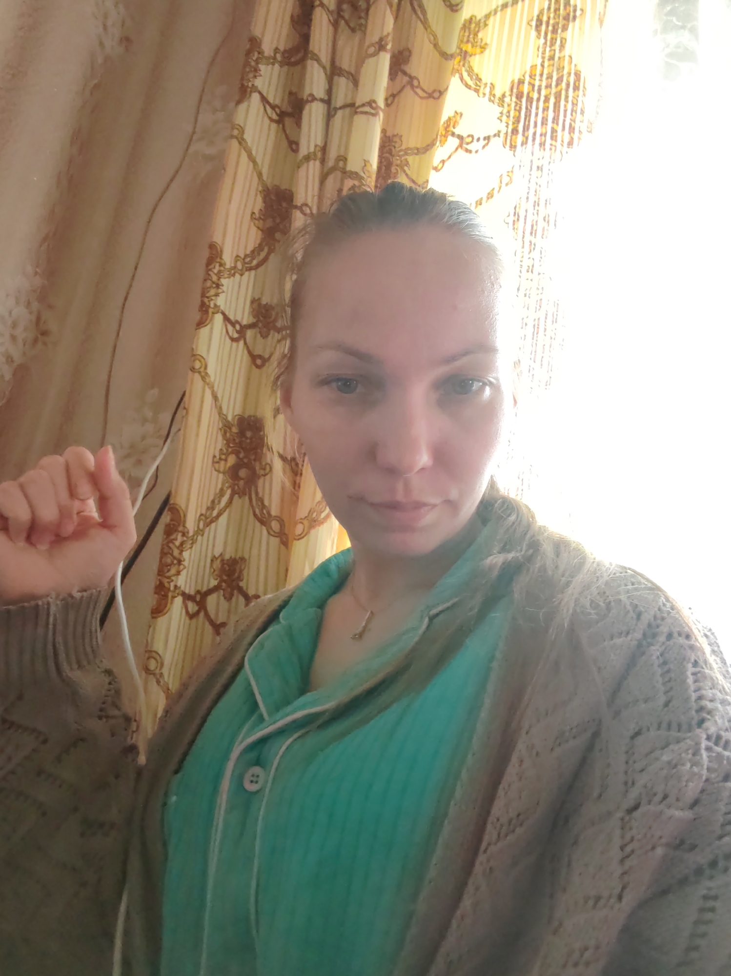 A woman is captured in a close-up, chest-up shot indoors, likely in Чухлэм, Russia. She has her hair pulled back and looks directly at the camera with a neutral expression. She is wearing a teal-colored, button-up top with white piping, possibly a pajama top, layered with a light-colored, intricately knit or crocheted cardigan. A delicate necklace is visible around her neck. Her left hand is raised to shoulder height, with fingers slightly curled, appearing to be in the process of holding something or posing.
The background features patterned curtains. To the left, a plain beige curtain or wall section is visible. To the right, a window with very bright, overexposed light creates strong backlighting, obscuring much of the detail in that area of the background. The intense natural light suggests it is daytime, possibly morning or afternoon. There are no visible texts in the image.