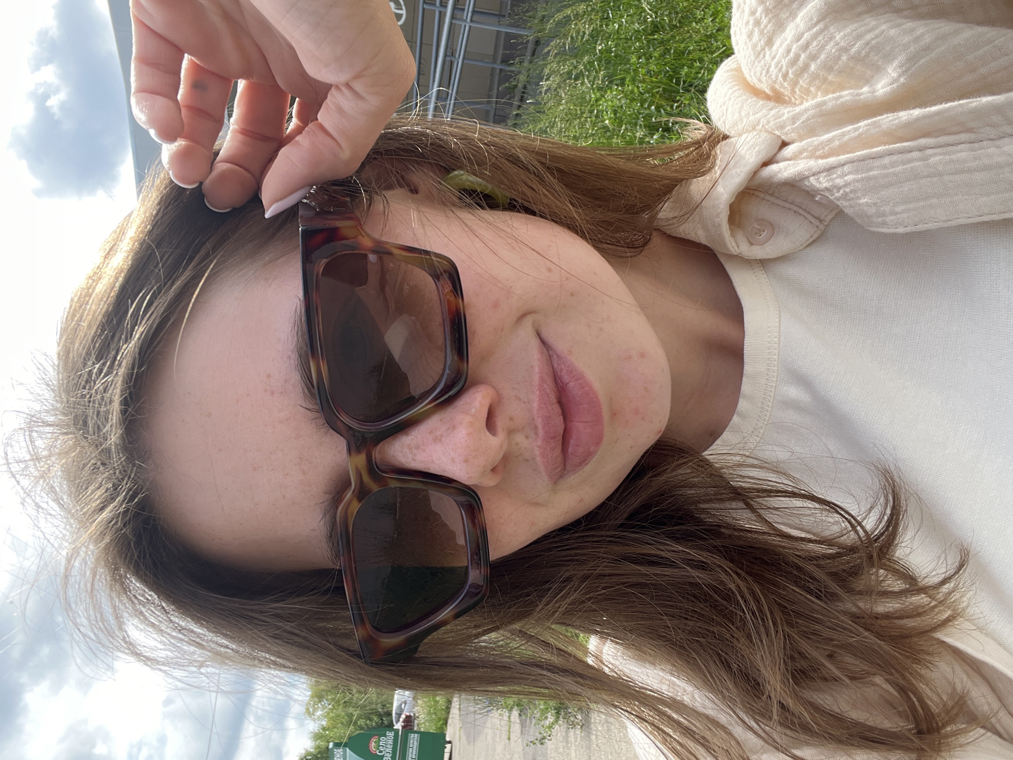 A young woman with light brown hair and freckles is captured in a selfie, outdoors. She is wearing large, tortoiseshell-patterned sunglasses and a light-colored, collared shirt or jacket over a cream-colored top. Her right hand is raised, adjusting her hair near the top of her head. Green, hoop-style earrings are visible. The background suggests an urban environment, with a hint of a green sign bearing Cyrillic text, possibly indicating a street name or advertisement, in the upper left. There is also evidence of grassy areas and a paved surface. The lighting indicates a daytime, possibly sunny, scene. The overall impression is casual and spontaneous. The location is specified as Chaikovsky, Russia.