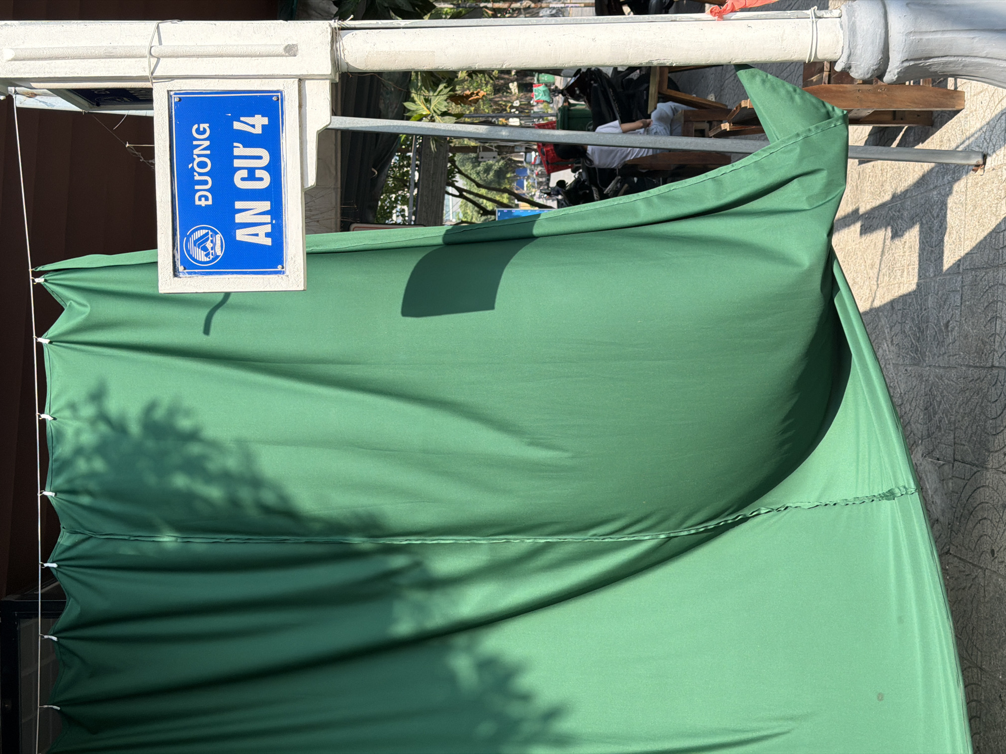 The image depicts a blue and white sign indicating "ĐƯỜNG AN CƯ 4" (An Cu 4 Road) in Đà Nẵng, Vietnam. A large, green canvas or awning is draped in the foreground. The sign is mounted on a white structure, possibly a building or support beam. Sunlight is visible, casting shadows on the canvas and nearby pavement. Some people and other objects can be seen in the distance, viewed through a gap above the green material. The foreground is partially obscured by the green canvas.