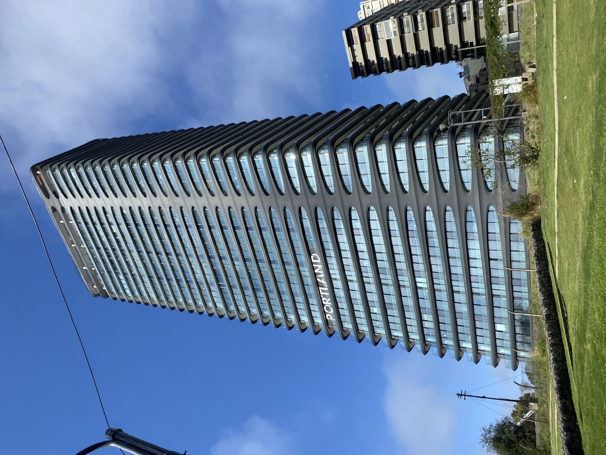 The image shows the Comega building in Buenos Aires, Argentina. It is a tall, modern building with a unique design. The building has rounded edges on the facade with oval windows. The word "PORTLAND" is written on the side of the building. A smaller building is also visible in the background, along with grass and trees. The sky is blue with some clouds. A utility pole is also visible.