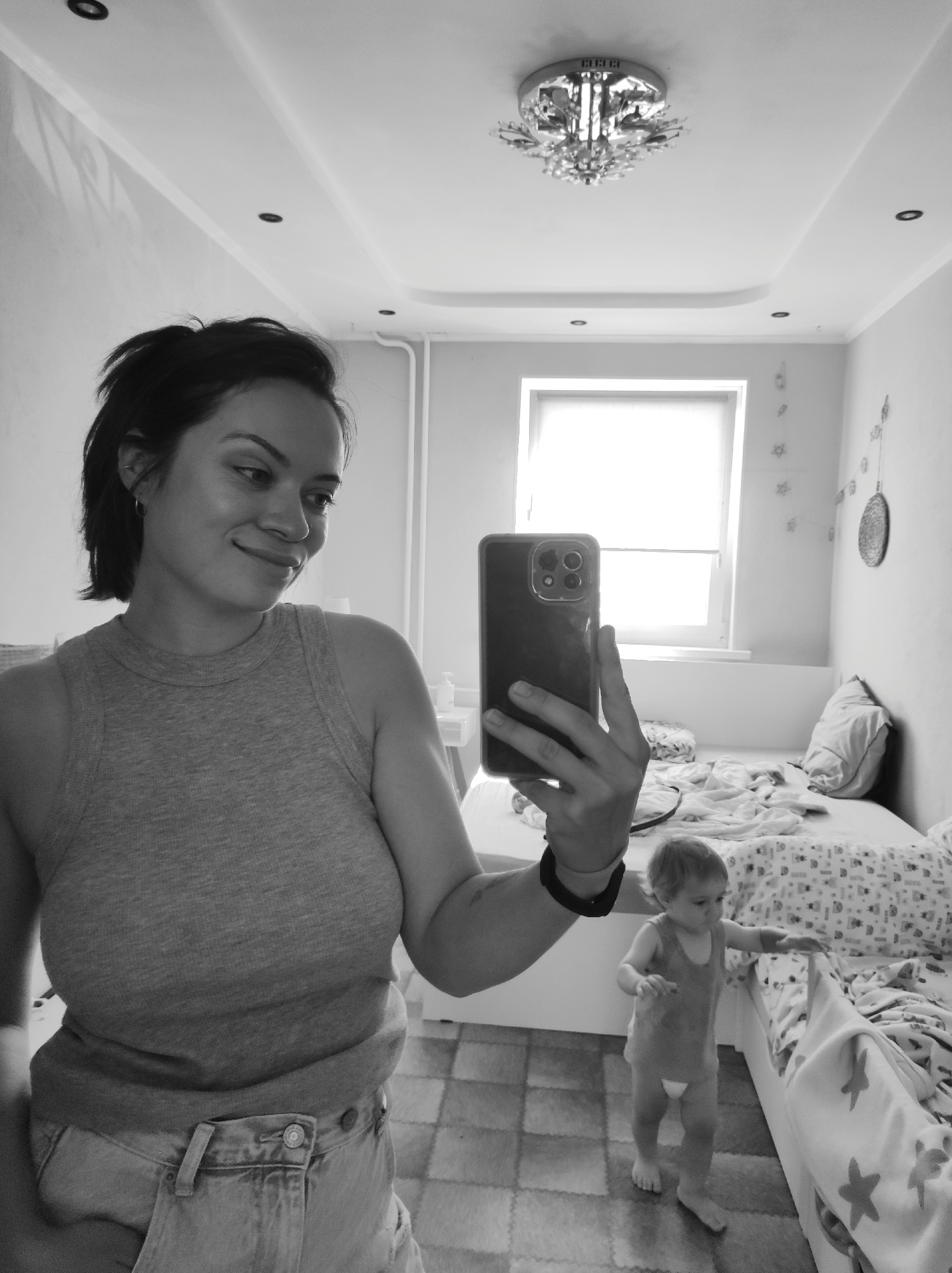 A grayscale, full-body, indoor, mirror selfie shows a woman holding a smartphone in her right hand, taking the photo. She is wearing a grey tank top and denim shorts. She has dark, short hair styled to one side and is looking to her right with a slight smile. The mirror reflects the room behind her, which appears to be a child's bedroom. In the background, a toddler stands on a tiled floor next to a bed covered with a patterned duvet. The room features a window with blinds, a ceiling light fixture, and decorations on the wall. The location is identified as Chaikovsky, Russia.