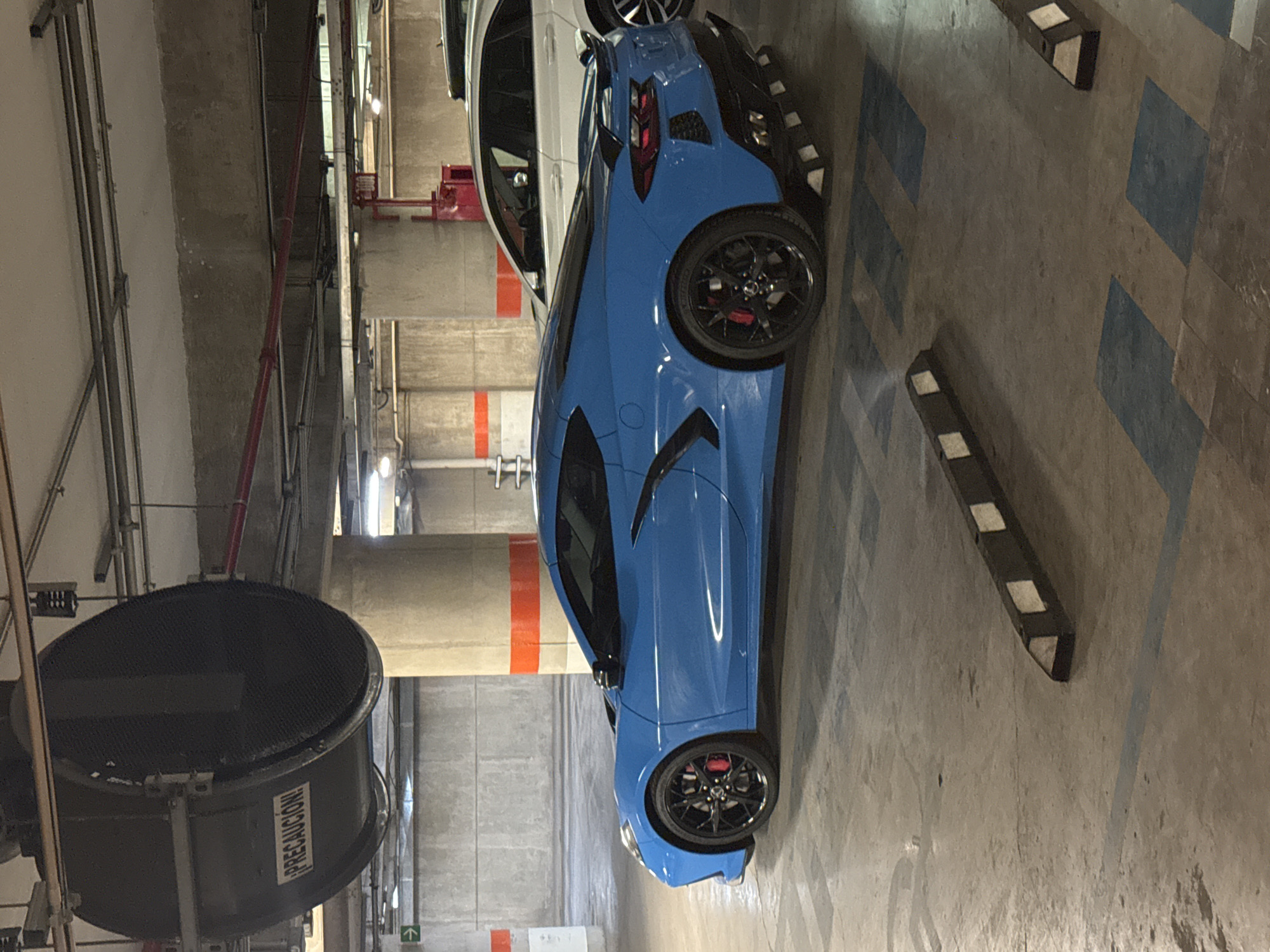 The image depicts a blue Chevrolet Corvette parked in a parking garage. Next to it, a white car is parked, partially visible. The garage has concrete columns with orange stripes and a large, industrial ventilation fan with the word "PRECAUCION" on it. Parking bumpers are present on the ground, which is marked with blue paint for parking spots. The scene is a parking garage, although the exact location in San Bernardino Tlaxcalancingo, Mexico, is not discernable from the image.