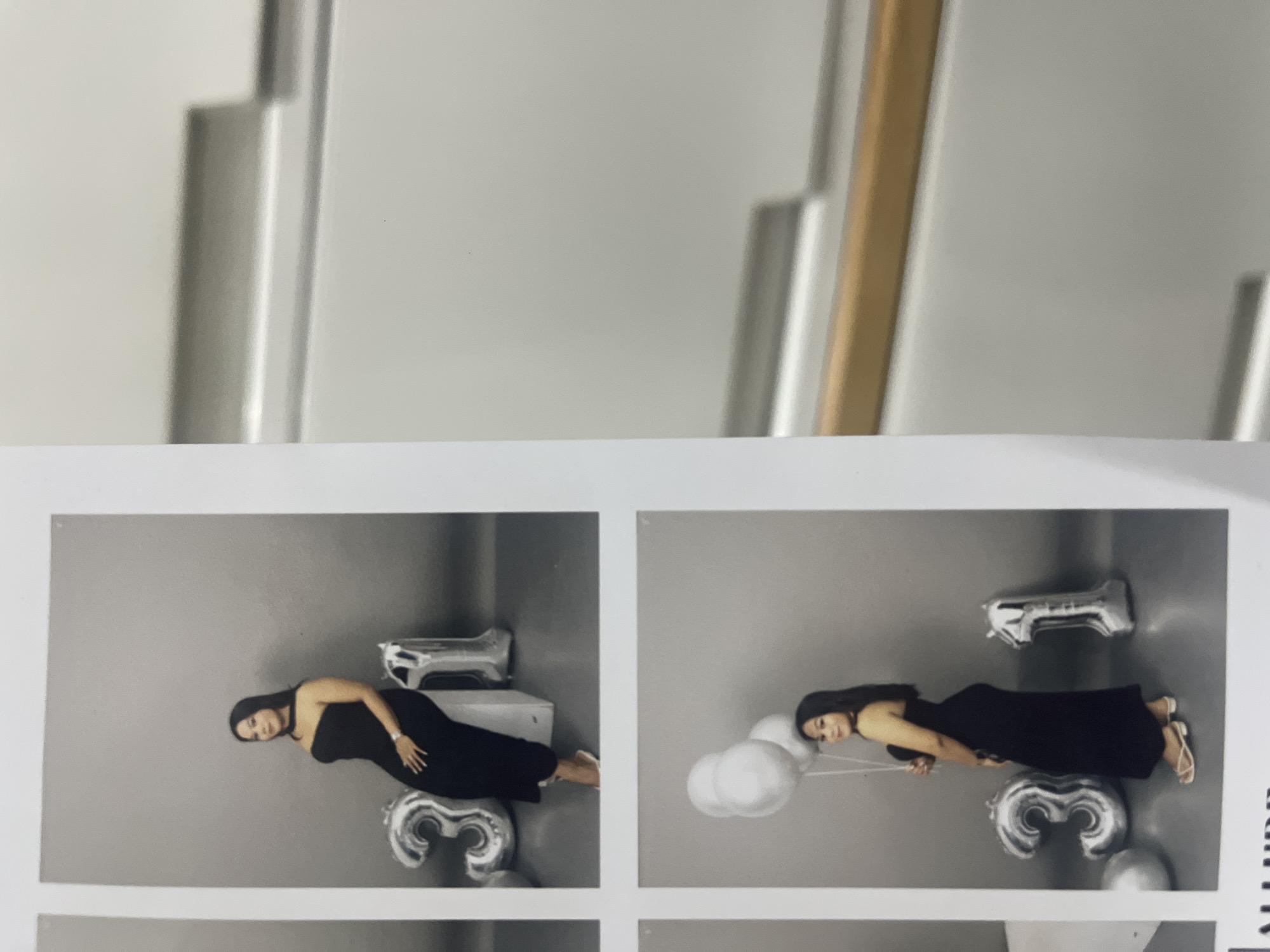 The image displays a portrait session featuring a woman. She is wearing a long, black, strapless dress and light-colored sandals. In the left panel, she is positioned horizontally against a grey backdrop, with large, silver foil balloons shaped like the numbers "2" and "3" placed near her. A silver rectangular object is also visible above her. In the right panel, she is again posed horizontally, this time holding a string attached to several white, cloud-shaped balloons. A silver foil balloon shaped like the number "3" is visible below her. The lighting appears to be from a studio setting, suggesting an indoor photoshoot. The woman has a neutral or slightly smiling expression. The context suggests this is a photoshoot for a special occasion, possibly a birthday, given the presence of the number balloons. The provided location context is Denpasar, Indonesia, but no specific geographical cues are visible within the image itself.