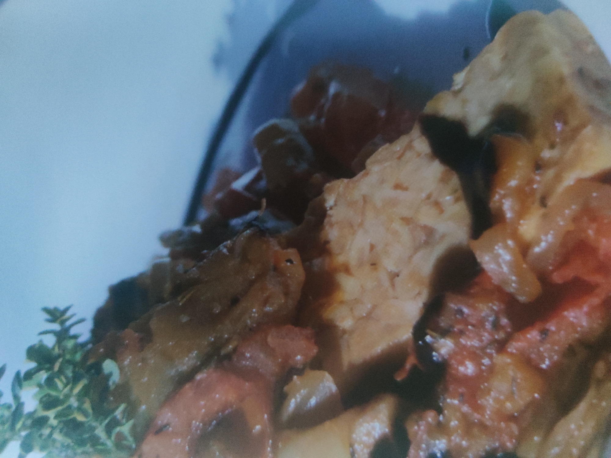 A close-up food photograph shows a dish with what appears to be tofu or tempeh pieces in a sauce, with some tomato and possibly mushrooms or other vegetables. There is also a sprig of thyme visible at the lower left of the image. The scene appears to be indoors, potentially in a home or restaurant setting. The city is Unknown, Unknown.