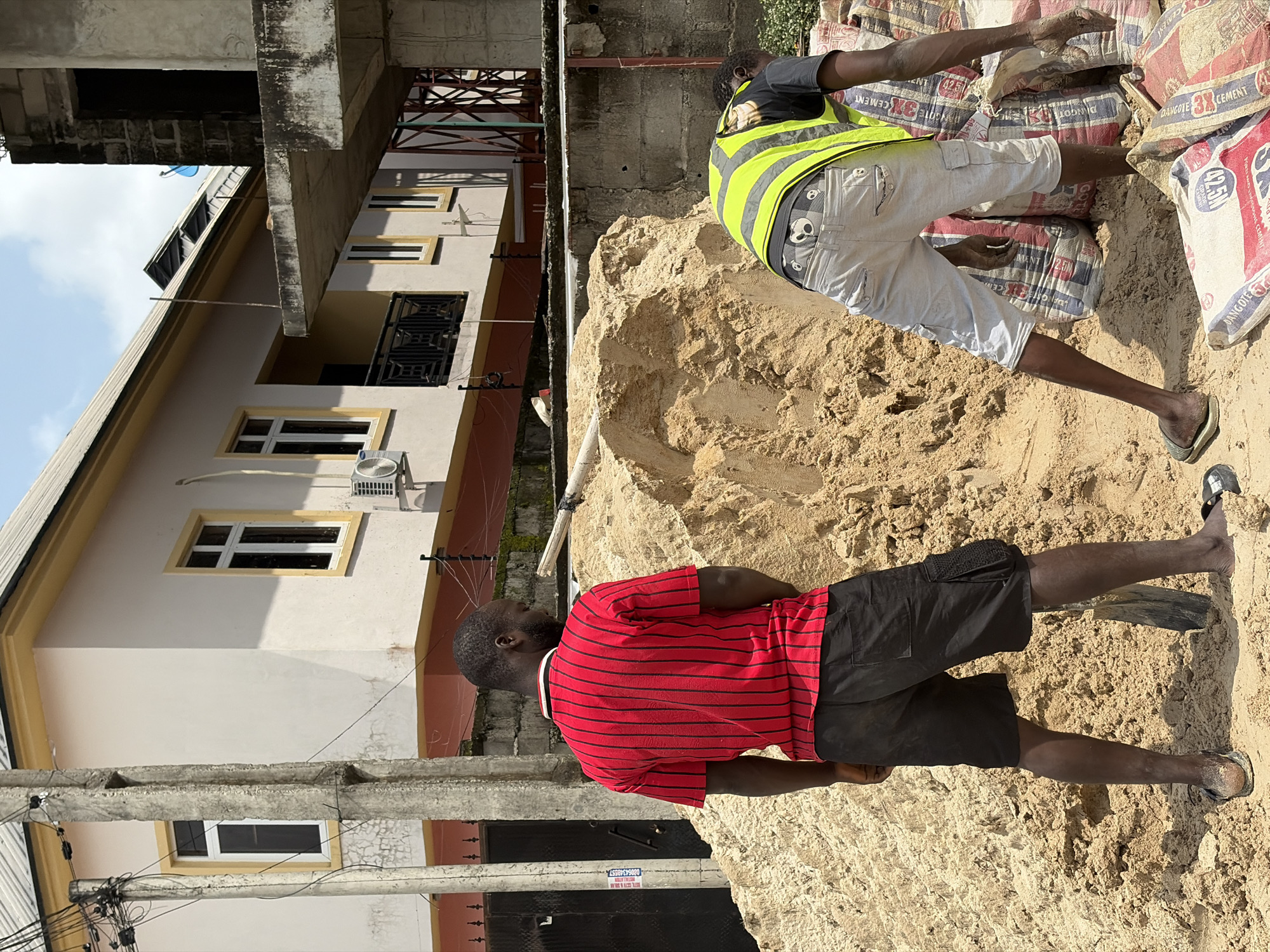 This media file depicts an outdoor construction scene in Igboefon, Nigeria. Two men are present, both appearing to be construction workers. The man in the foreground is wearing a red and black striped t-shirt and black shorts, with black sandals. He is standing on a large pile of sand. The second man is in the background, wearing a bright yellow reflective vest over a t-shirt and light-colored shorts. He is also standing on the sand pile, near stacks of cement bags.
The activity appears to be construction or site preparation, with the sand and cement bags suggesting building work is underway. The setting includes a multi-story building with visible windows and balconies to the left of the frame. Electrical wires and conduits are also visible, indicating the presence of infrastructure. The weather appears to be sunny, casting shadows on the sand and buildings. The overall scene suggests a typical day on a construction site in a populated area, likely during daylight hours. Some text is visible on the cement bags, including "DAWCOTE 3X CEMENT" and "42.5N". There is also a sign with text that is partially legible, possibly a contact number or business information. No specific emotions are conveyed by the subjects, as their faces are not clearly visible or are turned away.