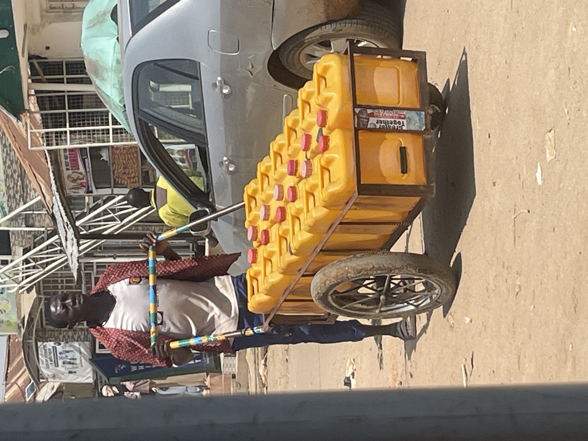 In an outdoor market setting in Abuja, Nigeria, a Black man is pushing a cart loaded with yellow jerrycans. He is wearing a white t-shirt with a red and black patterned shirt over it and dark blue trousers. In his hands, he holds a colorful painted bar, likely part of a mechanism for pushing or balancing the cart. The cart, made of metal, is filled with multiple yellow jerrycans, each topped with a red cap. A sticker with the words "Together" is visible on the back of the cart. Behind the man and the cart, a silver car is parked. In the background, there are market stalls with various items for sale and some overhead structures, suggesting a busy commercial area. The ground is sandy and dusty, characteristic of many urban environments in Nigeria.