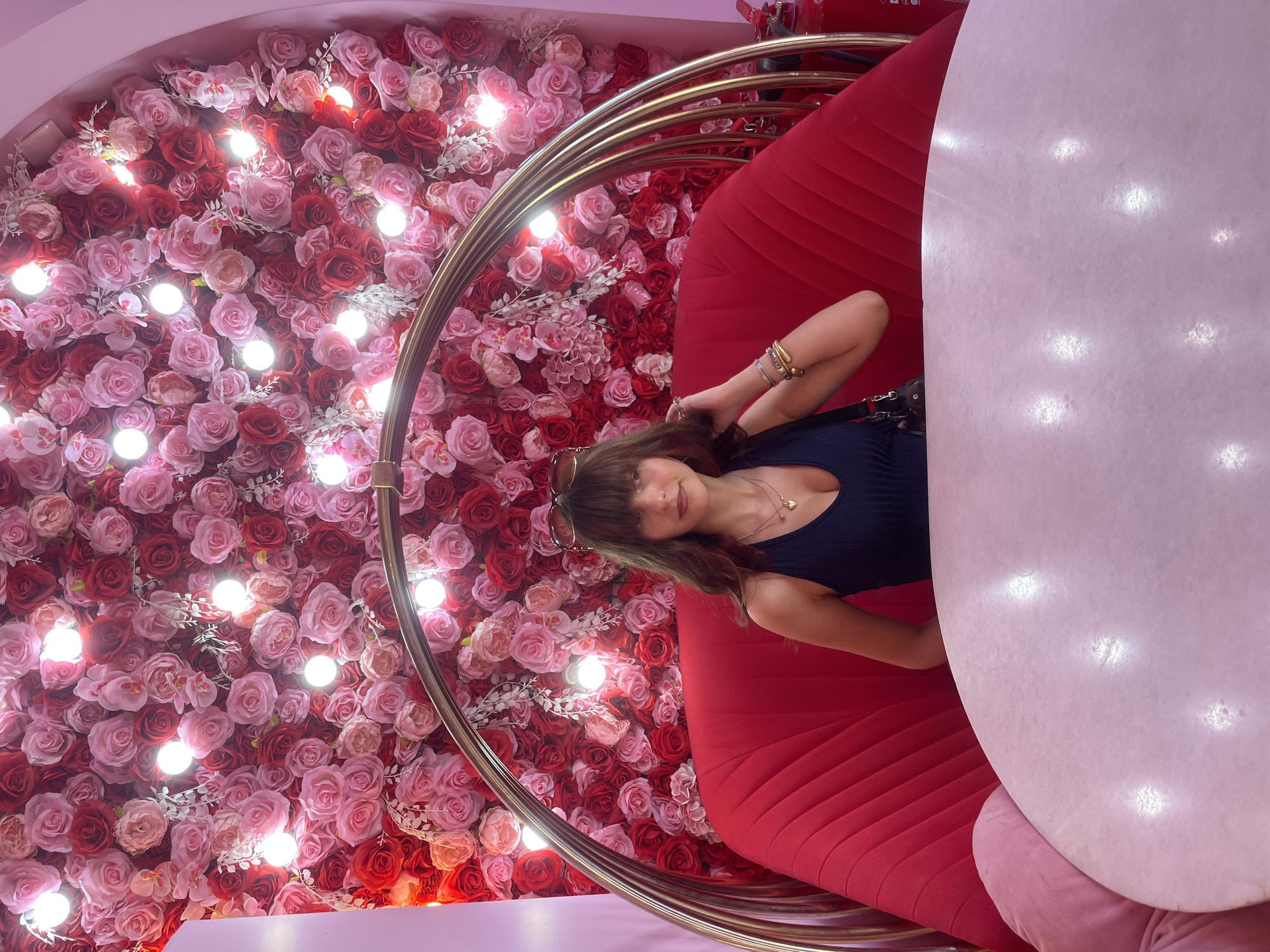 This media file captures a young woman posing for a photo in a vibrantly decorated setting. She is seated on a plush red sofa, which is part of a unique, lip-shaped chair with a golden frame. The backdrop is a wall densely covered with artificial pink and red roses, interspersed with small, bright lights, creating a romantic and festive atmosphere.
The woman has long brown hair and is wearing a dark blue sleeveless top and a heart-shaped necklace. She is also accessorized with several bracelets on her left wrist and is holding her hair with her right hand, while looking towards the camera with a subtle smile. Sunglasses are perched on her head.
To her right, a large, rounded pink surface, possibly a table or another seating element, dominates the frame, its surface reflecting the scattered lights. The overall impression is one of a whimsical and possibly commercial location, perhaps a themed cafe or a photoshoot studio, designed for visually appealing content. The lighting suggests it's indoors, possibly during the day or evening, with artificial lighting playing a significant role in the ambiance. The image conveys a sense of playful engagement with the environment. There is no visible text in the media file.