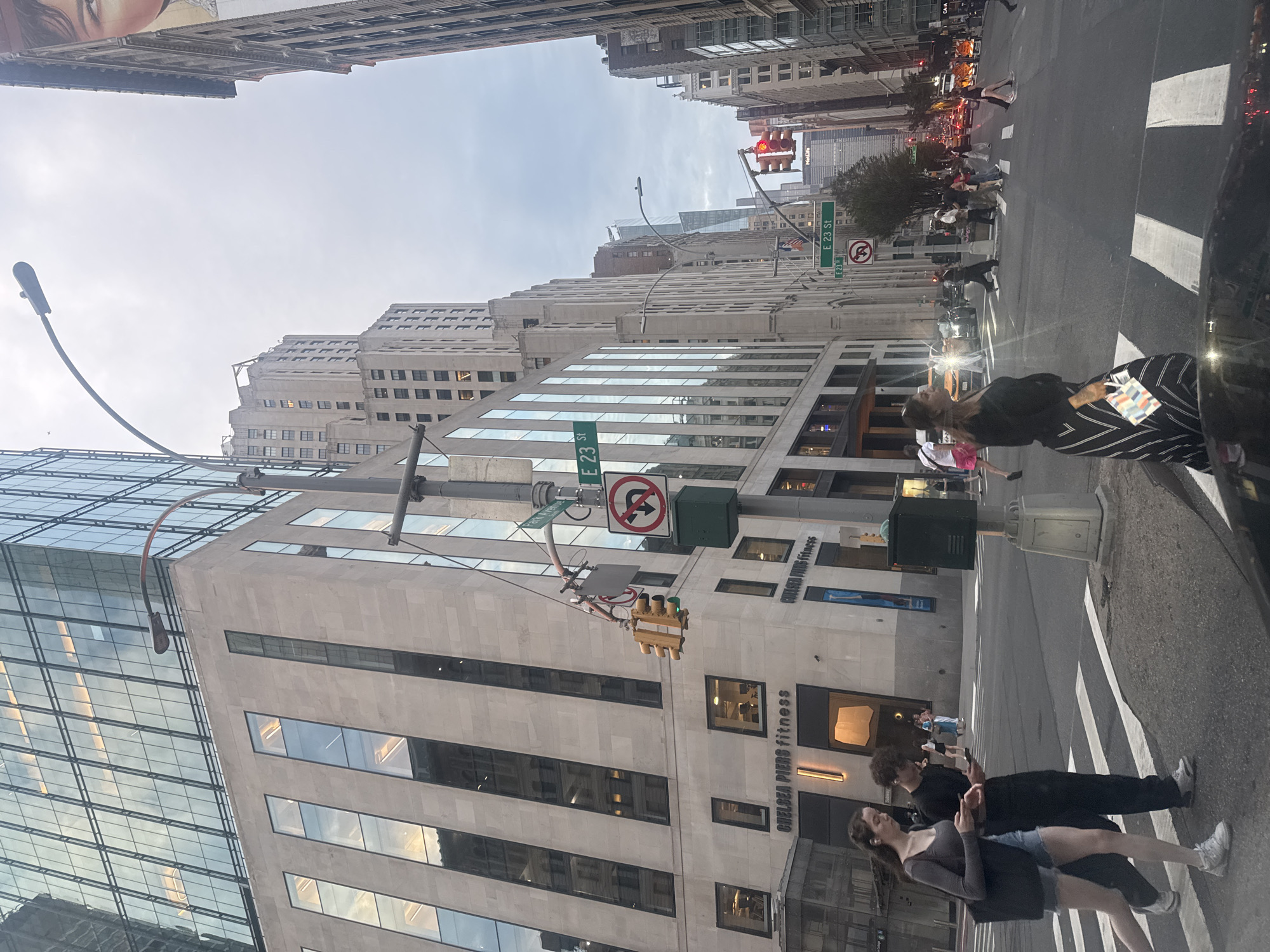 This image captures a street scene in New York City, likely Manhattan, with a view down an avenue lined with tall buildings. In the foreground, a street corner with pedestrian crosswalks is visible. Two young adults, a female with brown hair wearing a grey long-sleeved shirt and denim shorts, and a male with dark curly hair wearing a black t-shirt and black pants, are walking across the street, both looking at their phones. To the right, a woman in a black and white striped skirt and black top walks towards the viewer. Traffic lights and street signs, including "E 23 St," are visible, indicating the location. Several cars and taxis are present in the street, suggesting active urban traffic. The architecture of the buildings ranges from modern glass-fronted structures to older, more ornate buildings. The sky appears overcast. The presence of "Chelsea Piers Fitness" signage on one of the buildings provides a specific landmark.