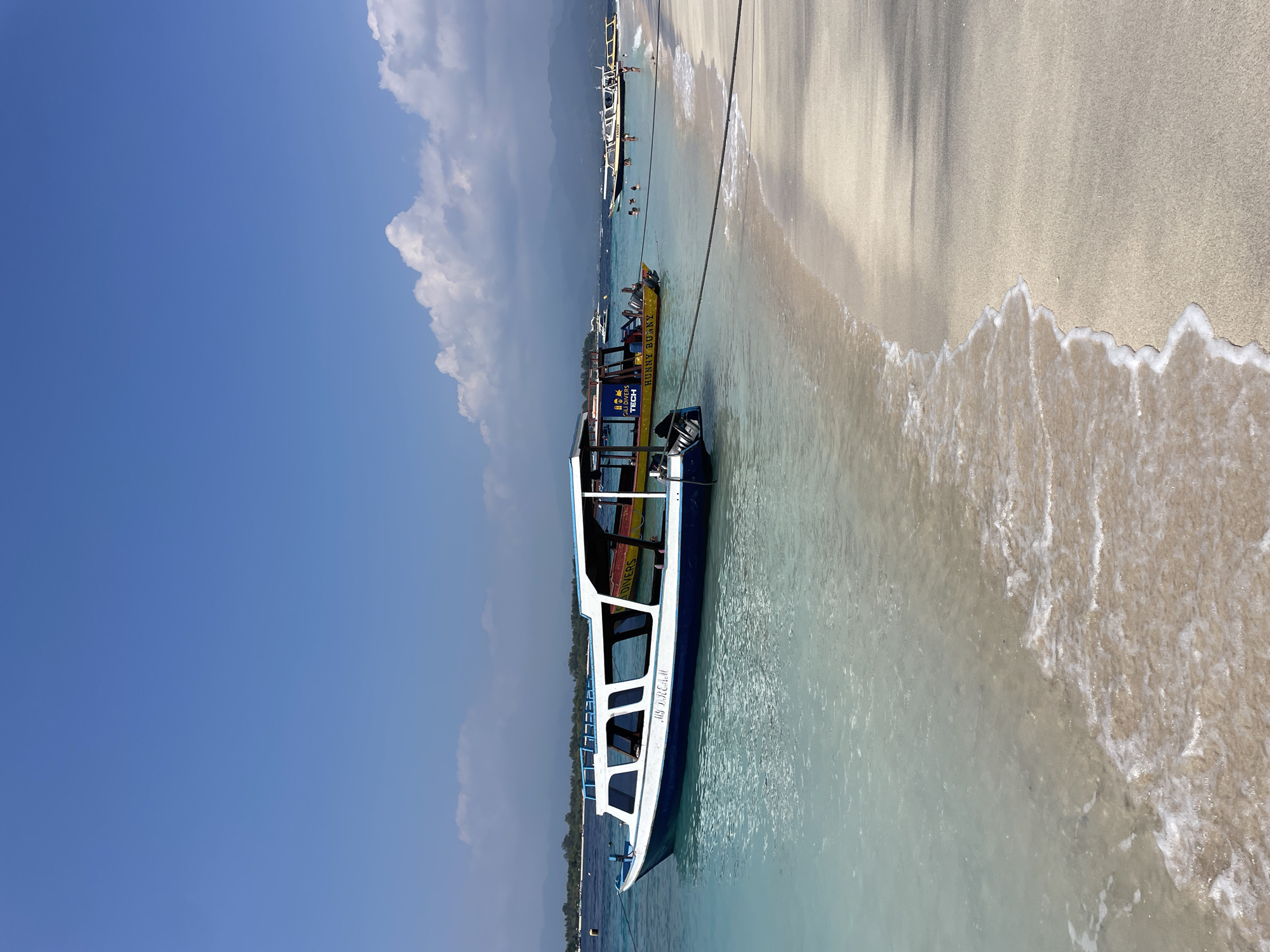 A scenic view of a tropical beach in Gili Trawangan, Indonesia features several boats docked near the shore. The foreground showcases the clear, turquoise waters lapping against a sandy beach, with waves gently breaking on the sand. In the mid-ground, a white and blue boat with a covered seating area is prominently displayed, its name "MY DREAM" visible on its side. Behind it, a yellow boat with the name "HUNNY BUNNY" is also moored. Several other boats are visible further out in the ocean. The background reveals a hazy view of distant islands or mountains under a bright blue sky with scattered white clouds. The scene depicts a tranquil and idyllic beach environment.
