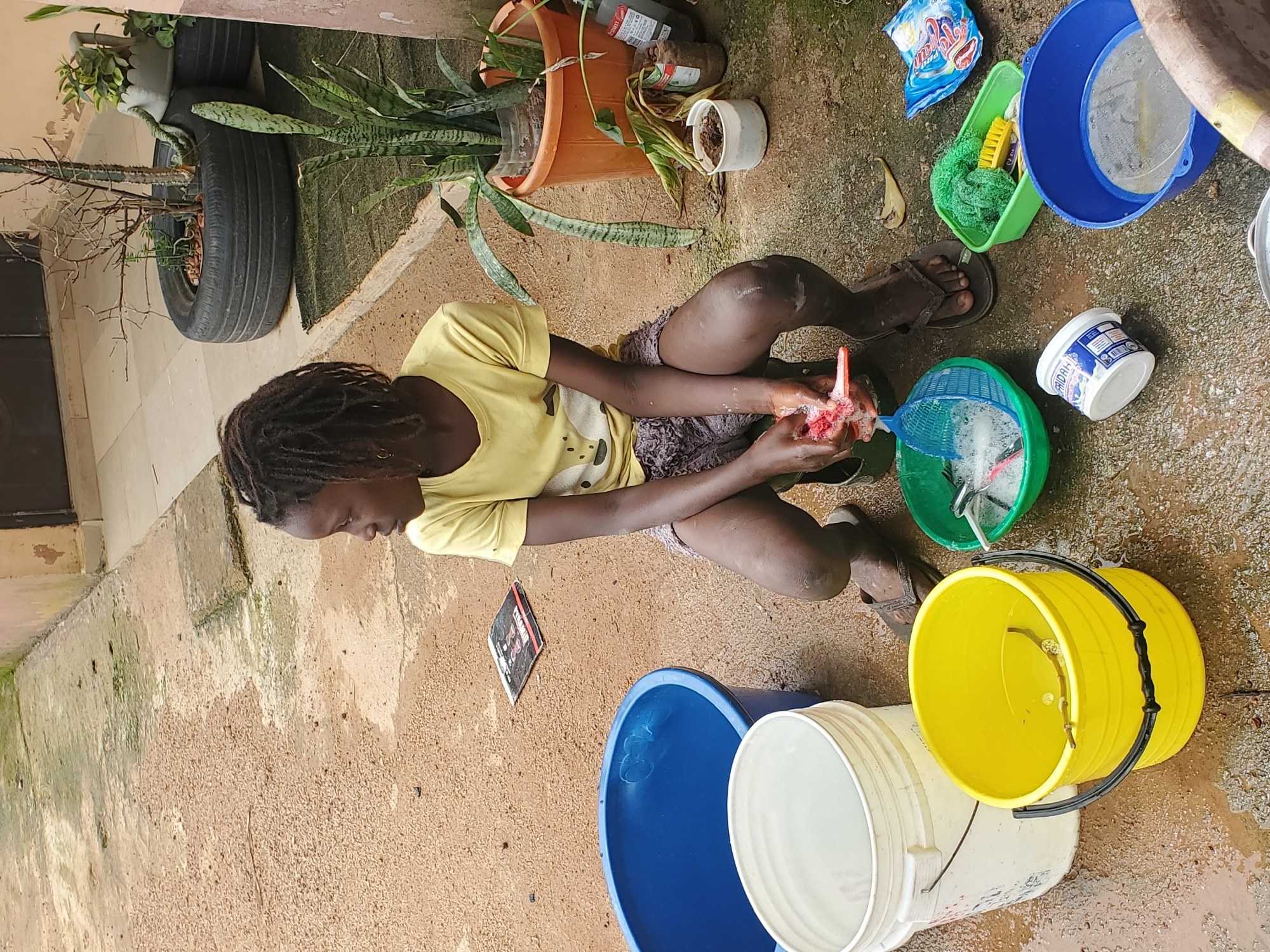 In Rafin Bauna, Nigeria, a young Black person with dreadlocks, wearing a yellow t-shirt and dark shorts, is crouching on a wet, sandy ground next to several buckets and cleaning supplies. The person is actively engaged in washing something, with their hands in a green colander filled with soapy water and a red object, possibly a fish or food item.
Surrounding the person are various containers: a large blue bucket, a white bucket, and a yellow bucket containing water and a stick. Other items present include a green plastic tub with a yellow brush, a blue bowl with a sieve, a white tub of what appears to be cream or soap, and a blue packet of detergent.
The environment suggests an outdoor area, possibly a yard or communal washing space, with a building visible in the background and several potted plants, one in a repurposed tire. The ground is uneven and speckled with dirt and some scattered debris. The overall scene depicts a task being performed outdoors, likely related to food preparation or cleaning, in a somewhat rustic setting.