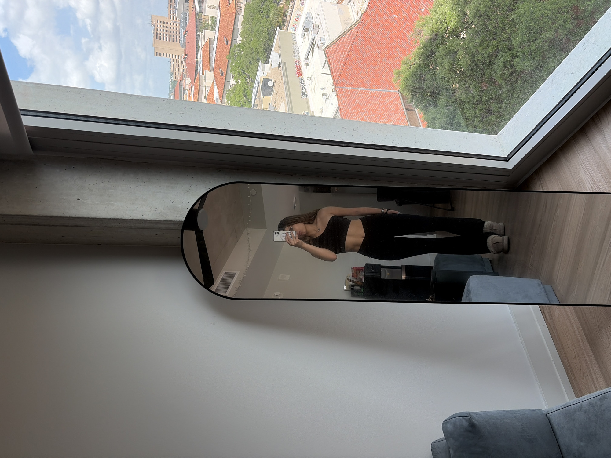 A vertical, full-shot photograph shows a woman taking a mirror selfie in an apartment setting in Austin, Texas, USA. The woman, reflected in the mirror, is wearing a dark athletic top and black pants, holding a cell phone up to capture her image. She stands near a low, dark media console and a blue upholstered chair. Behind her, the window offers a view of Austin's cityscape, characterized by red-tiled roofs, dense green trees, and various building heights. The room's interior is primarily white, with hardwood floors visible in the reflection. The mirror is a full-length style with a rounded top and a black frame.