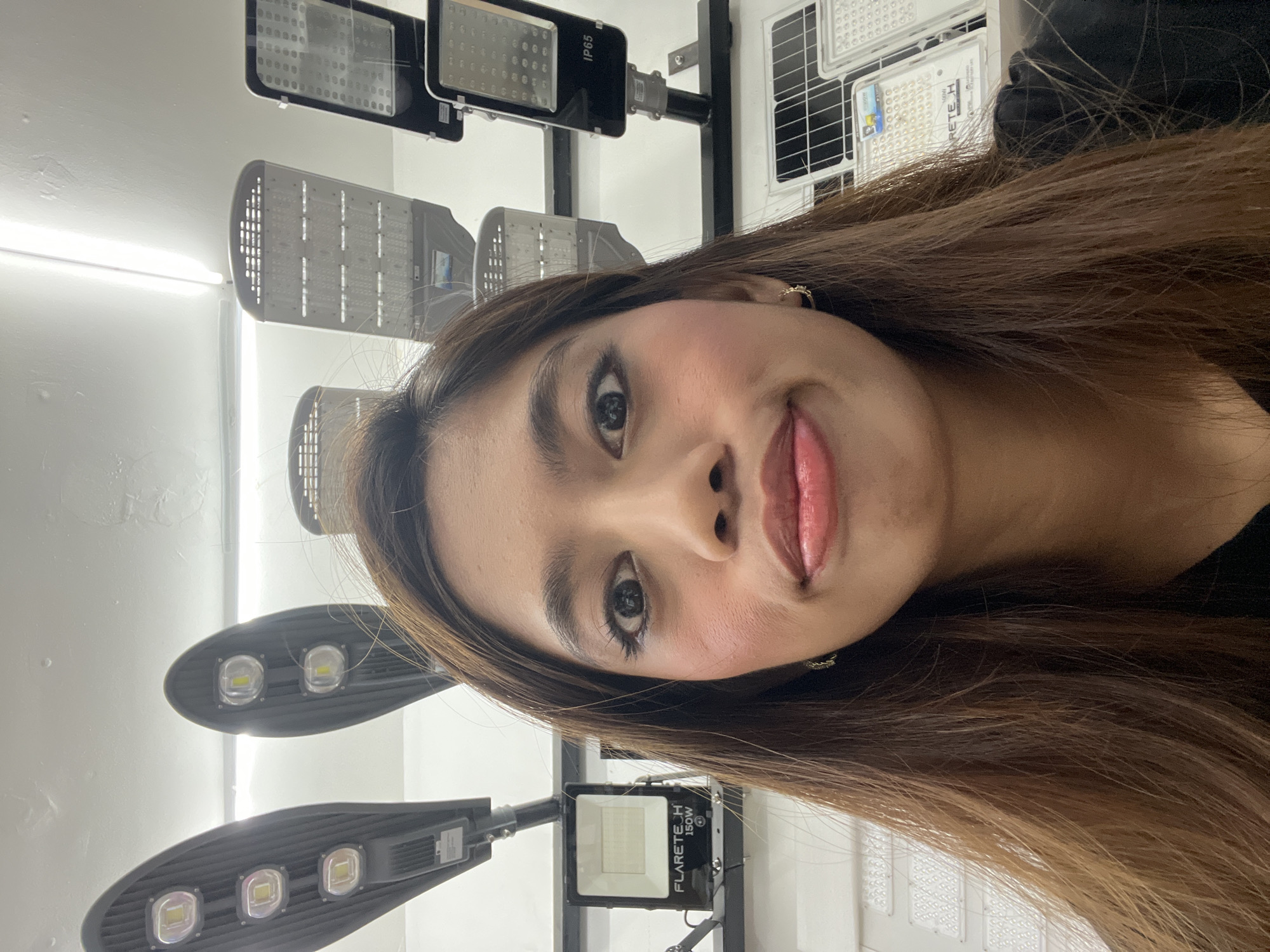 A young woman is the central subject of this indoor portrait, likely taken in a commercial setting in Davao City, Philippines. She has long, straight brown hair and is wearing makeup, including visible eyeliner and blush. Her lips are slightly parted, and she has a subtle, playful expression, looking directly at the camera with her head tilted. A small gold-colored earring is visible in her right ear, and she is wearing a dark top.
The background consists of various types of LED lights displayed on stands against a white wall, suggesting a lighting store, showroom, or a display area for electrical products. Several modern LED street lights with multiple individual LED chips are visible, arranged vertically. One of these street lights has "IP65" printed on its casing. In the lower right background, a rectangular LED floodlight is clearly seen, labeled with the brand "FLARETECH" and "150W". Other smaller, grid-patterned light fixtures are also visible further back. The overall lighting in the scene is bright, highlighting both the subject and the displayed products.