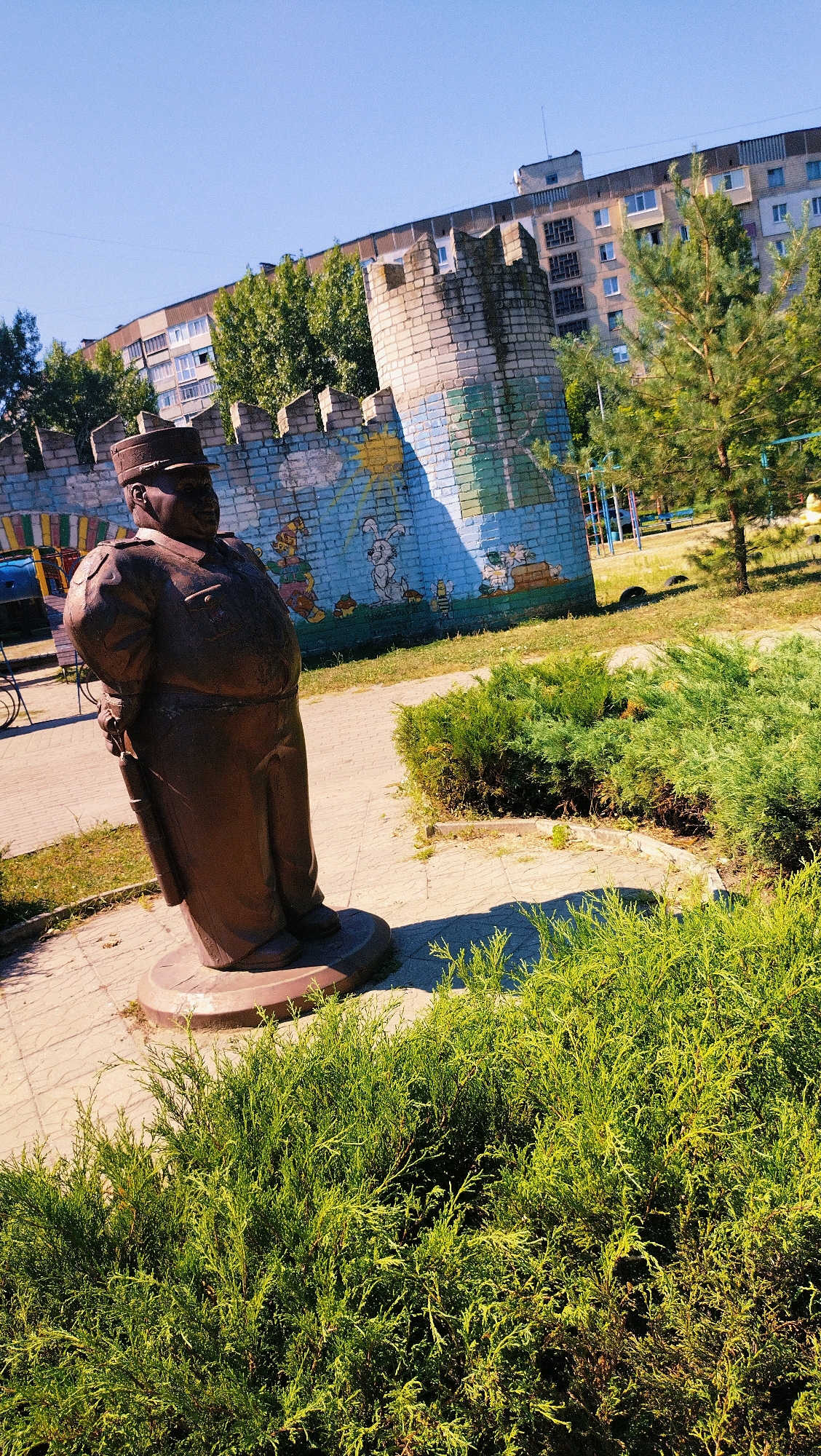 In a park-like setting under a clear blue sky, a bronze statue of a stout man in uniform, possibly a police officer or military personnel, stands on a paved circular base. He is depicted wearing a hat and a jacket, with his hands clasped behind his back, and a holster visible on his left hip. Behind the statue, a low, decorative brick wall with battlements painted with cartoonish figures of rabbits and other animals forms a backdrop. Beyond the wall, a multi-story residential building and trees are visible. The ground in the foreground is covered with lush green bushes and a paved pathway. The scene appears to be a public area, likely a playground or recreational space, in an urban environment.