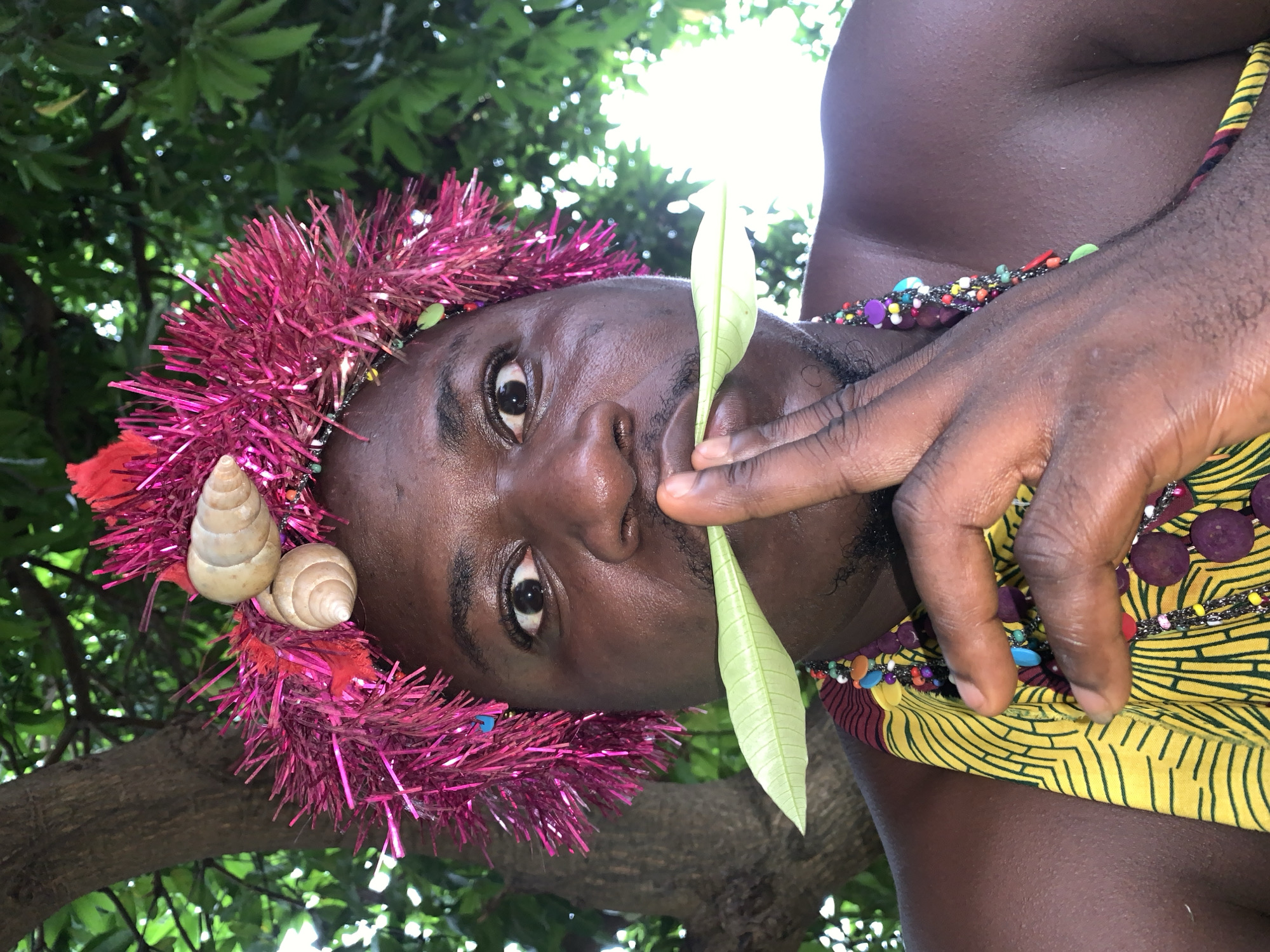 A close-up, low-angle, eye-level outdoor shot captures a dark-skinned person wearing a vibrant pink, tinsel-like headdress adorned with two light-colored seashells and red embellishments. The person's face is partially obscured by their right hand, which is holding a single, light-green leaf horizontally across their mouth. They are looking directly at the camera with wide eyes, and a portion of a dark, multi-strand beaded necklace is visible against their shoulder and chest. The person is wearing a yellow garment with a repeating dark green abstract pattern. The background is dominated by the blurred green foliage of trees and branches, suggesting an outdoor environment, potentially in a natural setting within or near the city of Akwanga, Nigeria. The lighting indicates a bright, daytime setting, possibly with dappled sunlight filtering through the leaves.