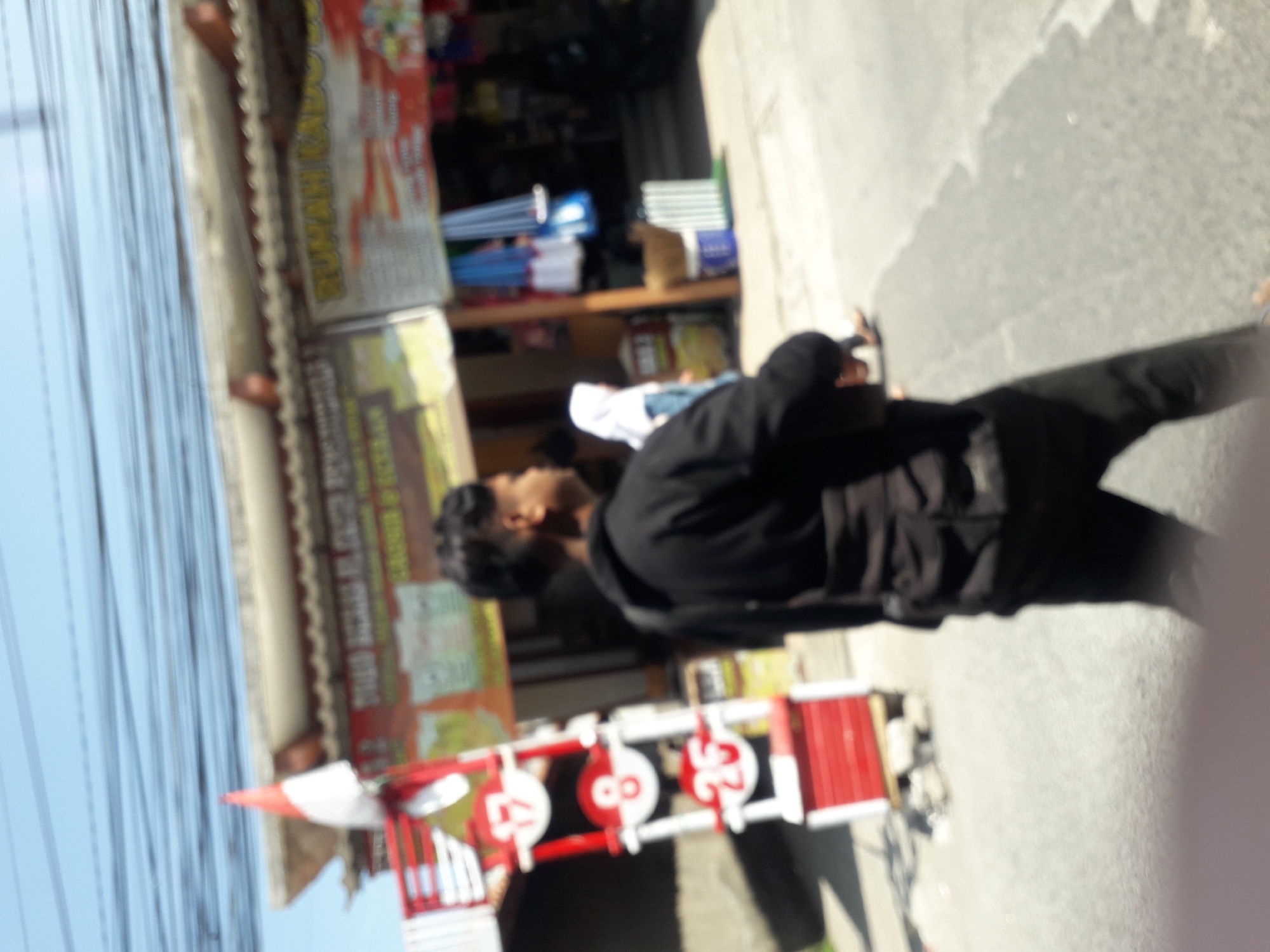 A blurred, outdoor, daytime image captures a street scene in what is identified as Linggar, Indonesia. In the foreground, a young male wearing a black hoodie and dark pants is walking away from the camera, his left shoulder visible. He has a black bag slung across his chest. To the left of the frame, a red and white structure with several circular elements displaying numbers "17", "8", and "25" is partially visible. Behind the young man, a female wearing a white hijab and light blue clothing is walking. Further back, a storefront with awnings and various merchandise is visible. The sky is clear and blue, with power lines stretching across the top of the frame. The overall impression is a street view with pedestrians and commercial activity under bright daylight.