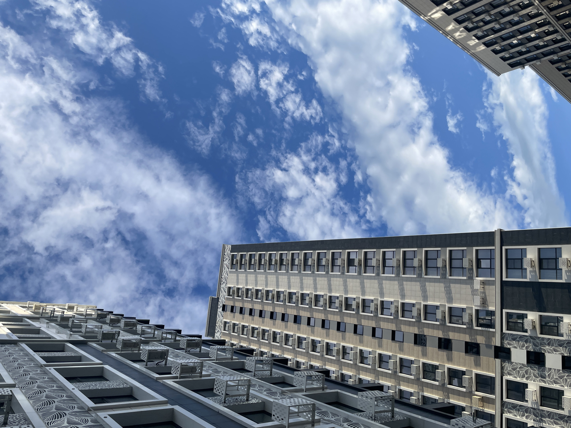 The image shows a low-angle view of modern high-rise apartment buildings against a bright blue sky with scattered white clouds. The buildings feature a contemporary architectural design with numerous windows and balconies. On the left, a portion of one building is visible, showcasing a facade with intricate patterns and a series of external air conditioning units. To the right and further back, another taller building dominates the center of the frame, also displaying a grid of windows and external units. In the lower right corner, a third building section is partially visible, with a lighter colored facade and dark windows. The scene appears to be in an urban setting, likely a residential area in Stavropol, Russia, given the architectural style and context. There are no people or visible activity in the image.