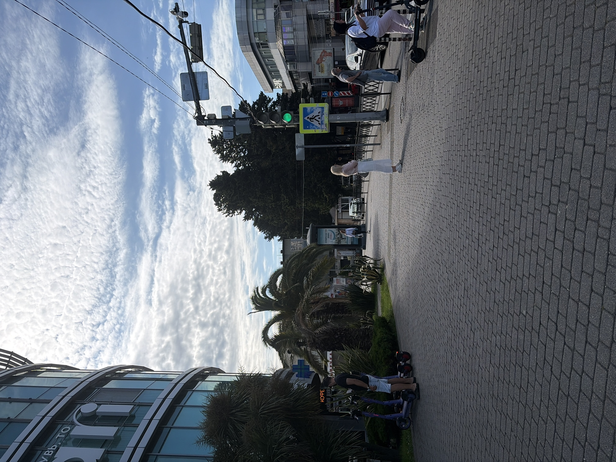The image depicts a street scene in Sochi, Russia, characterized by a modern building on the left and a sidewalk lined with trees and palm trees. Several electric scooters are parked on the left side of the walkway, with two individuals standing nearby. Further down the sidewalk, a pedestrian crossing is visible with a traffic light displaying green. Three people are standing at the crossing, and one is riding an electric scooter past them. Cars and buildings are visible in the background. The sky is filled with white, wispy clouds against a blue backdrop.