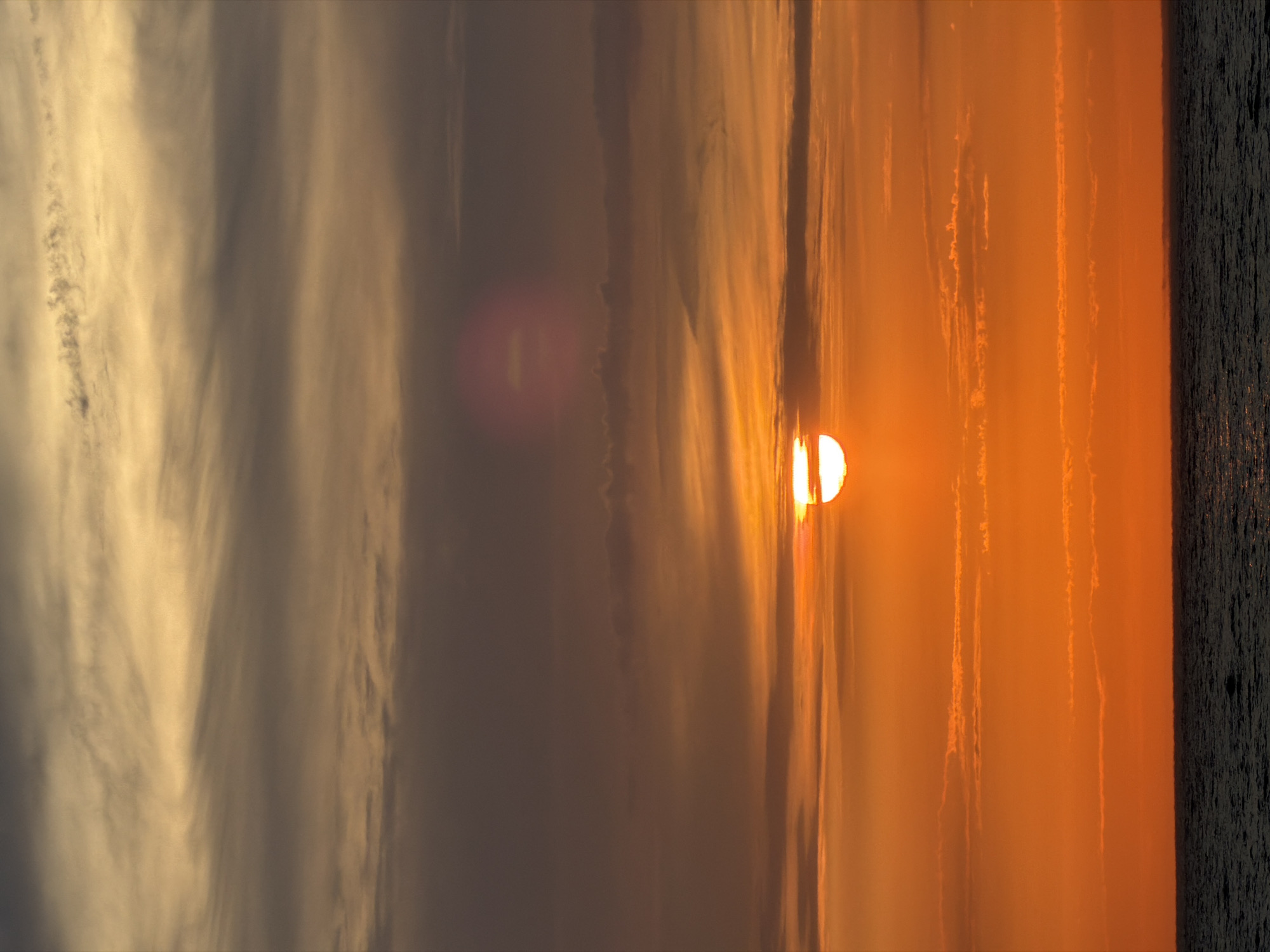 The image depicts a sunset over the Black Sea, likely near the city of Sochi, Russia. The sun, a bright white disc, is partially obscured by wispy clouds, casting an intense orange glow across the sky. Layers of clouds, ranging in color from dark gray to golden yellow, stretch horizontally across the frame. Below the horizon, the dark, choppy surface of the sea is visible. There are no people or man-made structures in the image, creating a serene and natural landscape. The overall impression is one of a dramatic and colorful end to the day.