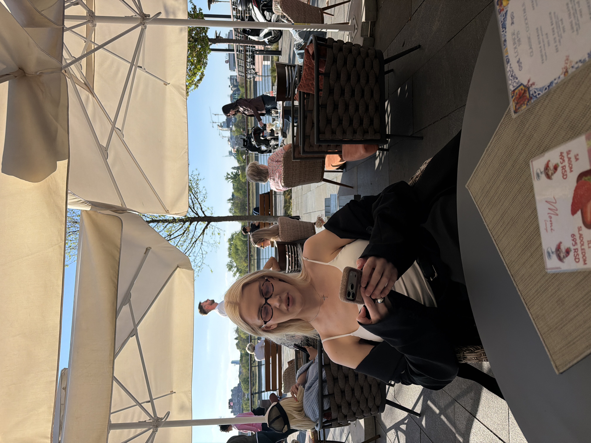 The image shows a woman with blonde hair and glasses sitting at an outdoor cafe in Belgrade, Serbia. She is holding a smartphone. The cafe is located on a waterfront, with other patrons and boats visible in the background. Tables are set up with umbrellas. The menu is on the table. The weather appears to be sunny and bright.