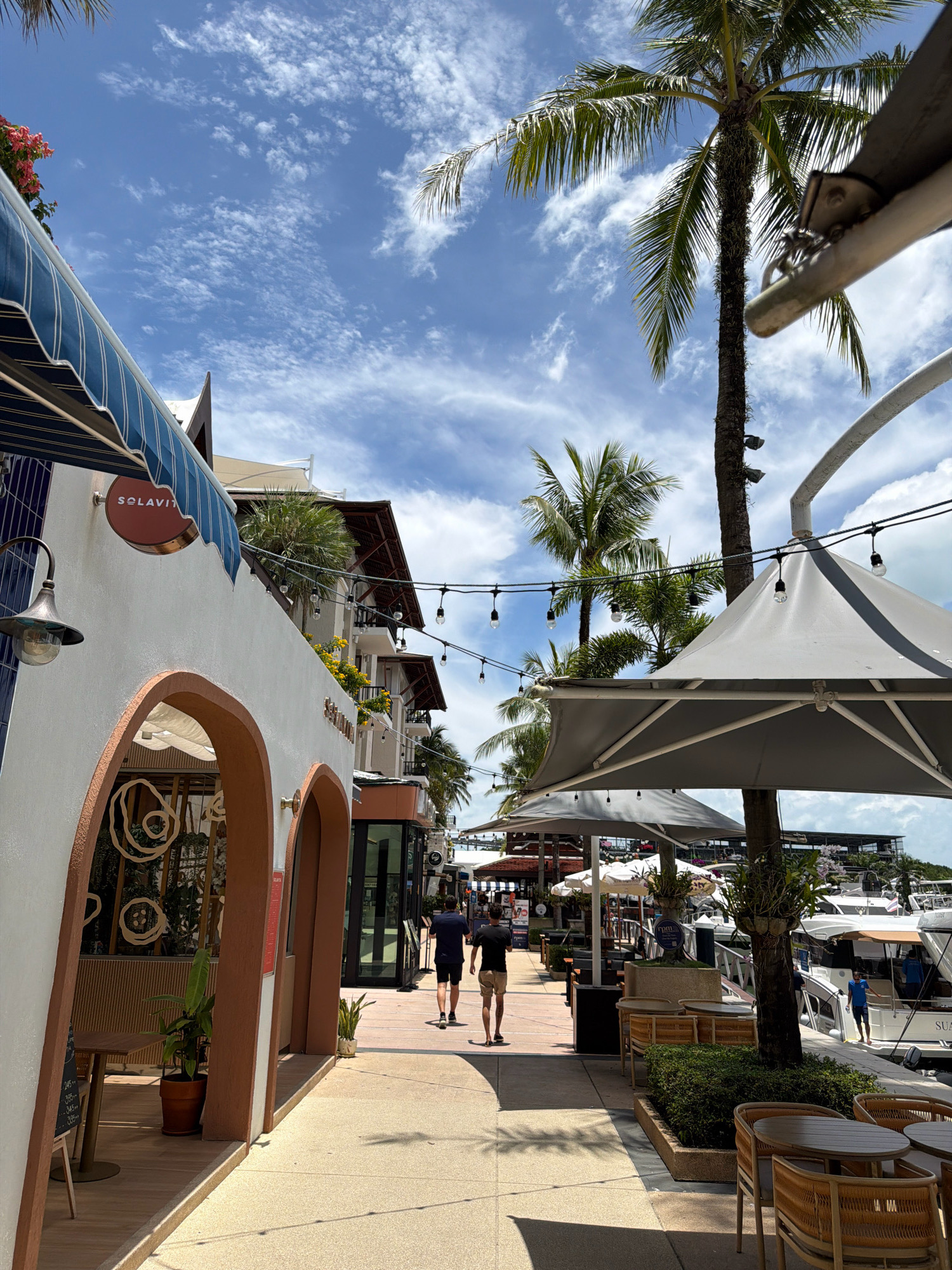 In the city of Ko Kaeo, Thailand, a pedestrian walkway lined with shops and palm trees leads towards a marina filled with boats. The walkway features an outdoor seating area with wicker chairs and tables, covered by large grey umbrellas. A man in a blue shirt and shorts is visible on a boat, while two men in dark shirts and light shorts walk away from the camera down the walkway. Buildings with blue awnings and arched entrances are situated on the left side of the walkway. The sky is blue with scattered white clouds, and the sun casts shadows on the ground.