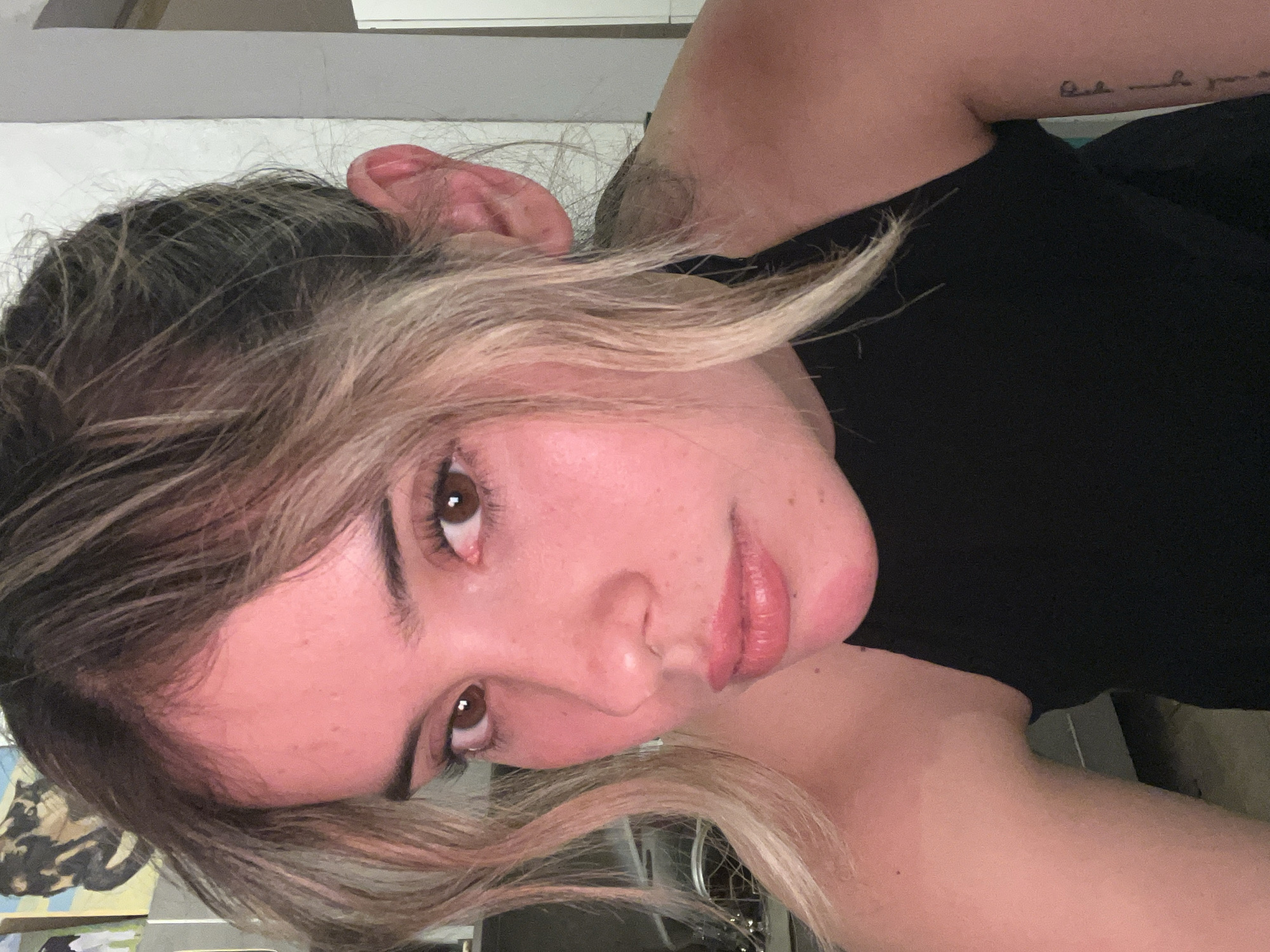 A close-up, waist-up photograph depicts a young Caucasian woman with blonde-brown hair styled in loose waves. Her skin is flushed pink, suggesting potential sunburn, particularly on her forehead and cheeks. She wears a black halter top. Her facial features include brown eyes, a small nose, and full lips. A partial view of a tattoo is visible on her right arm. The background is blurred, showing indistinct objects and a light-colored wall. The image quality is good, with clear focus on the subject. No specific location in Sumalao, Argentina, can be identified from the visual information provided.