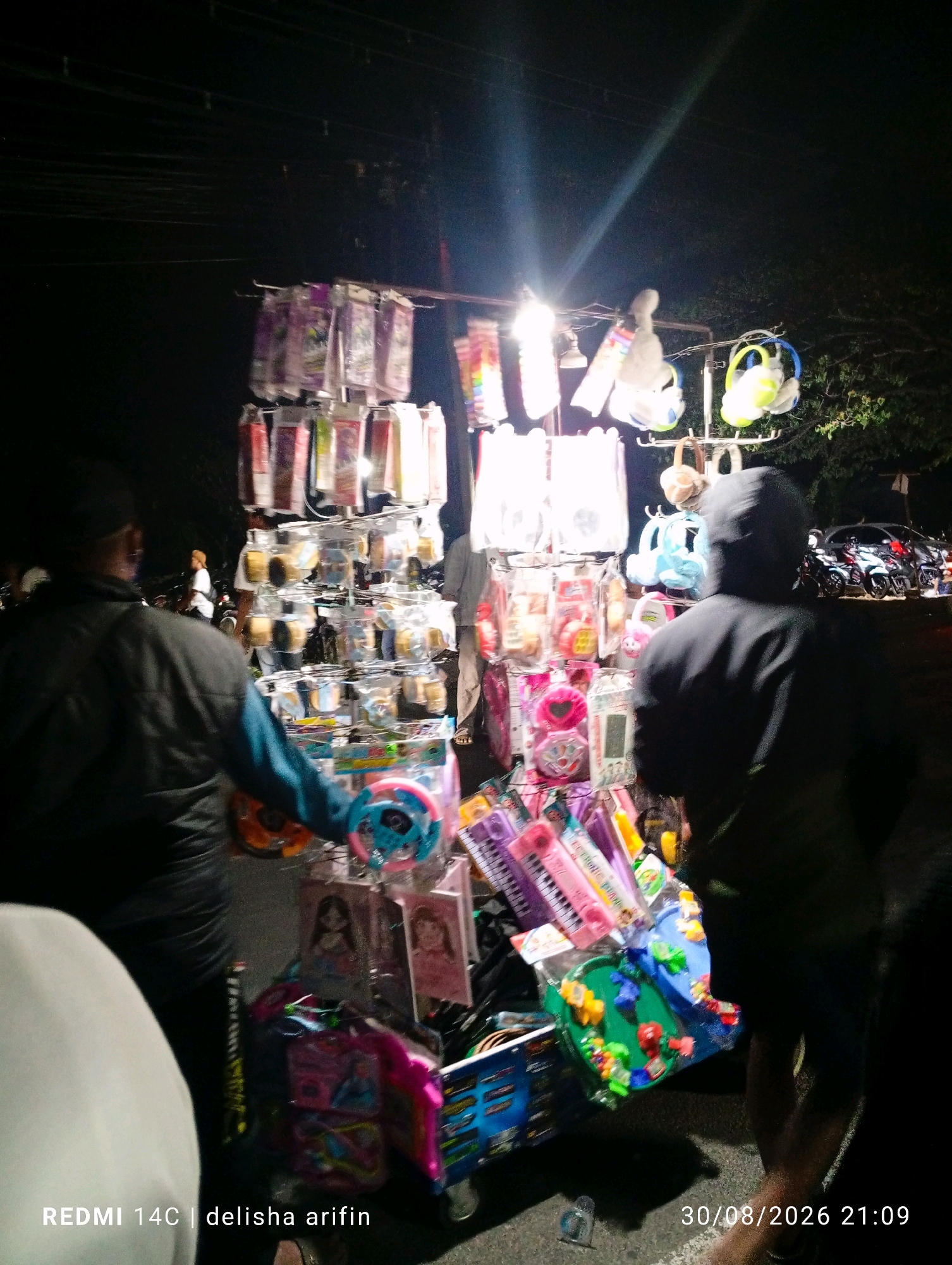 In Gambir Kuning, Indonesia, a street vendor is seen at night, displaying various items on a stall. The stall is laden with a variety of goods, including what appear to be toys, such as colorful spinning tops and plush toys hanging from racks. Packaged items, possibly stationery or small accessories, are also visible, arranged in rows on the display. Two individuals are present; one is a vendor looking towards the stall, while another, possibly a customer, is walking away from the camera, facing the other direction. The background reveals a dark, outdoor environment, with a few trees and parked motorcycles and cars suggesting a public street or marketplace. The lighting appears to be artificial, coming from the stall itself and potentially streetlights, casting shadows and creating a somewhat blurry, atmospheric scene. The timestamp "30/08/2026 21:09" is present in the lower right corner, and "REDMI 14C | delisha arifin" is in the lower left corner.