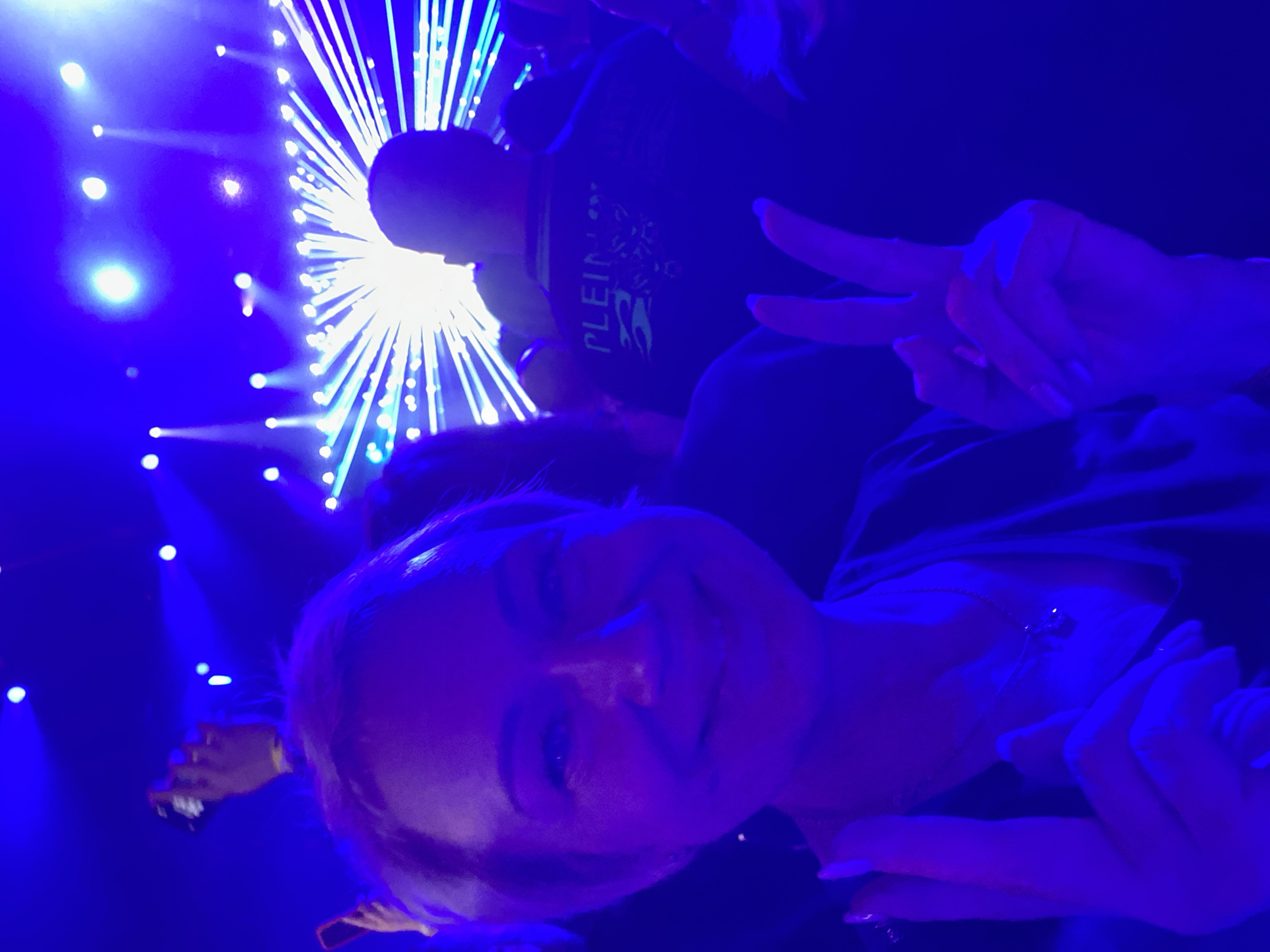 The image depicts a woman with blonde hair, smiling and making a peace sign with her right hand, as she takes a selfie at a concert. She is wearing a necklace. In the background, there is a crowd of people and a stage with bright blue and white lights, creating a dynamic light show. The lighting suggests it is nighttime, likely at an indoor venue, possibly a music hall or arena. The overall atmosphere is energetic and festive. The image does not contain any information or visual cues to suggest the location is in the city of Saint Petersburg, Russia.
