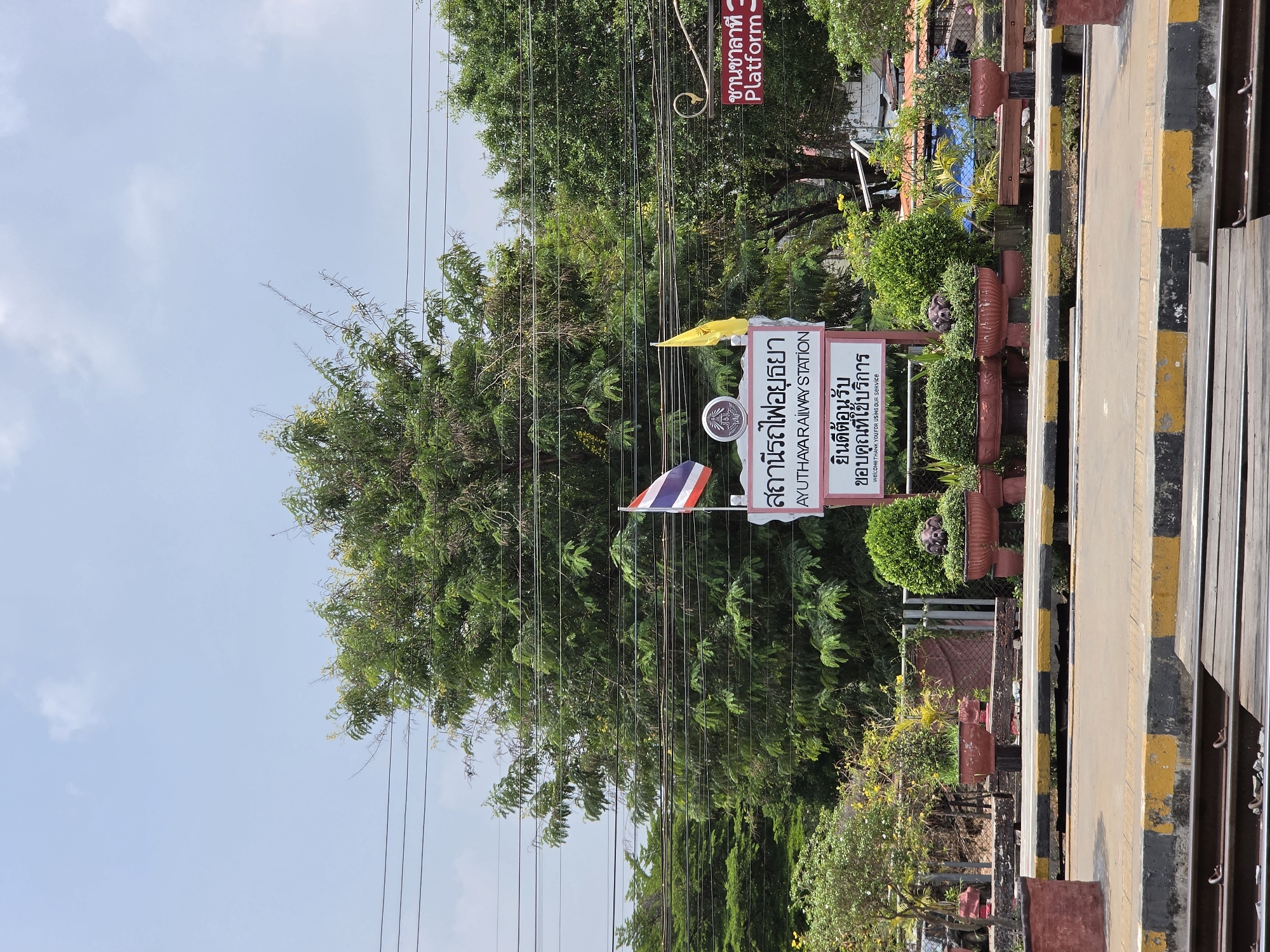 The image depicts the Ayutthaya Railway Station in Phra Nakhon Si Ayutthaya City Municipality, Thailand. A sign displays the station's name in Thai and English. The Thai flag and another flag fly beside the sign. Lush green trees surround the sign and station. A railway platform with yellow and black painted edges is visible. Power lines traverse the sky above the trees.