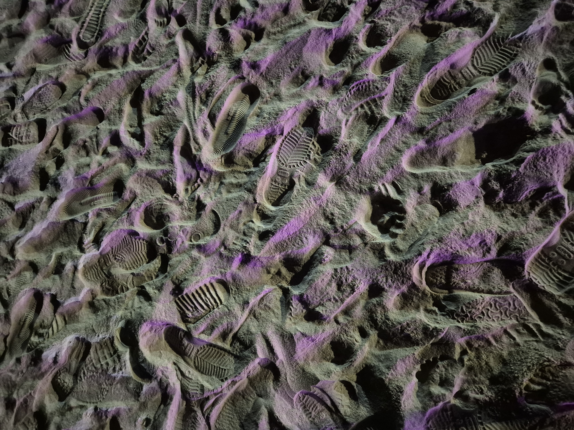 The image displays a dense field of shoe imprints in a sandy or dusty environment, illuminated with purple and green lighting. The imprints vary in depth and clarity, indicating a significant amount of foot traffic. Some imprints clearly show the tread patterns of different types of shoes, including what appear to be athletic shoes and boots. The ground surface is uneven, with raised and depressed areas, suggesting it is soft and malleable. The overall impression is of a heavily trafficked area where many individuals have left their mark. The specific location in Da Nang, Vietnam cannot be determined from the image alone.