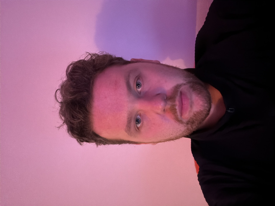 The image presents a head-and-shoulders portrait of a light-skinned adult male centered in the frame. He has dark, curly hair and a short, dark beard. His blue eyes are open and directed towards the viewer, and his expression is neutral. He is wearing a black collared top with a visible zipper partially open at the neckline. The background is a plain wall illuminated by a magenta-pink light source, creating a uniform purple-pink hue across the scene and reflecting strongly on the subject's face. A faint shadow is visible on the wall behind the subject's right side. The specific location within the city of London, United Kingdom, is not discernible from the visual evidence provided in the image.