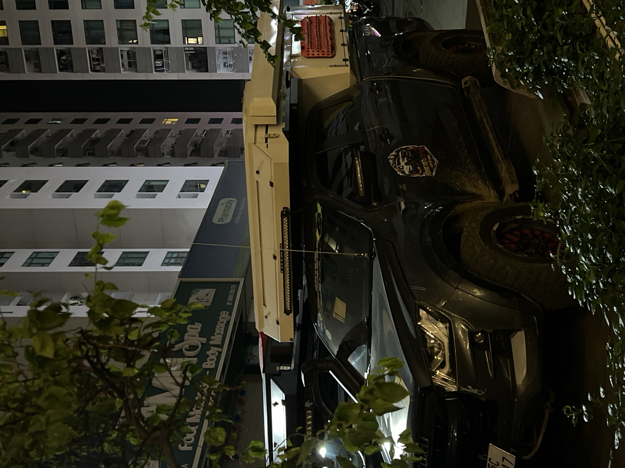 The image depicts a nighttime urban scene in Bac Nha Trang Ward, Vietnam, centered around a dark-colored, heavily modified pickup truck. The truck is equipped with a substantial, light-colored camper shell or overland rig on its bed, featuring a light bar above the cab and a red, textured recovery board mounted on its side. A spare tire is visible at the rear. A sticker on the truck reads "HOANG VIET" and "LIFE OF SONIAL", with a partially visible website address "WWW. HOANG VIET". Part of the license plate "SC 17" is also discernible.
In the background, modern, multi-story buildings line the street, their windows, some illuminated, suggesting residential or commercial occupancy. Street-level establishments include a sign for "Pharmacity" and a blue-green sign advertising "Spa, Body Massage" and "Foot M-", accompanied by a phone number: "0258 350 179".
Green foliage from trees and bushes partially frames the scene, appearing in the foreground and on the right side. The overall lighting indicates it is nighttime, with artificial illumination from buildings, streetlights, and the vehicle. The truck appears to be stationary, parked on what seems to be a city street or parking area. No individuals are visible in the image.
