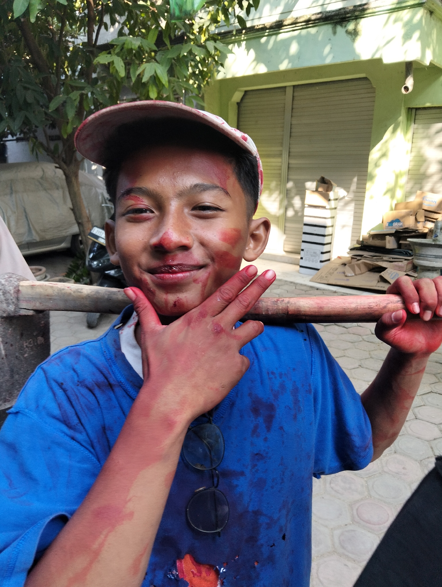 In Kranji, Indonesia, a young boy, likely a teenager, is captured in a close-up portrait. He wears a blue t-shirt and a red baseball cap tilted backward. His face and hands are smudged with red paint, suggesting he has been participating in an outdoor activity or event. He smiles at the camera, holding a wooden pole horizontally across his chest with both hands, and raising his left hand to his chin, showing off his paint-covered fingers. Behind him, a building with a green facade and rolling shutter doors is visible, along with trees and stacked cardboard boxes, indicating an urban or semi-urban setting. The lighting suggests it is daytime, with sunlight casting shadows.