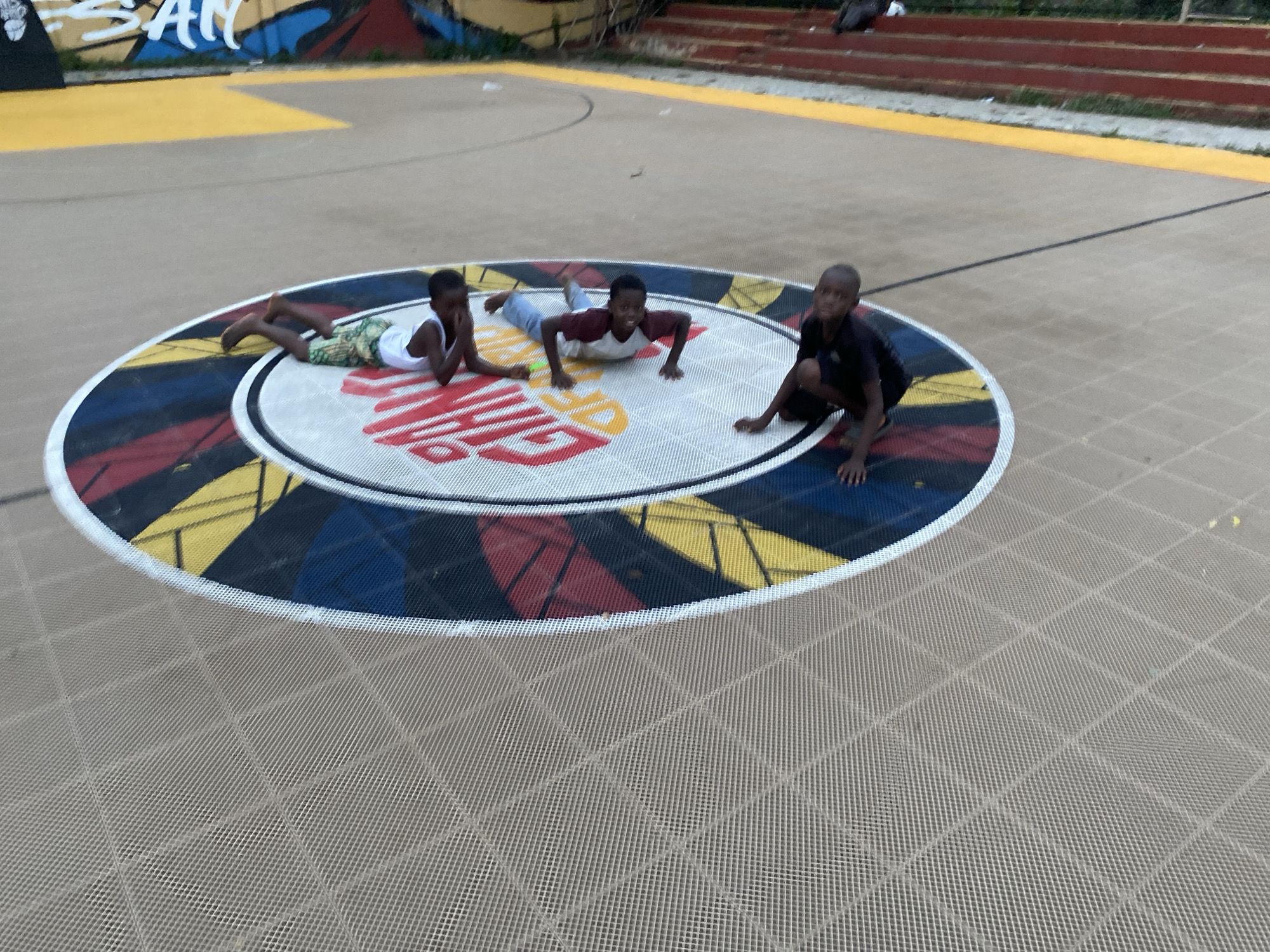 Three boys are positioned on a large circular design painted on a basketball court. The basketball court is located in Lagos, Nigeria.
The circular design features concentric rings of color: white, black, red, yellow, and blue. Inside the circle, "CITY OF LAGOS" is printed in large white letters. The court surface is a patterned tan material.
One boy is lying on his stomach. Another boy is in a push-up position. The third boy is sitting on the edge of the circle. Graffiti is visible on a wall in the background.