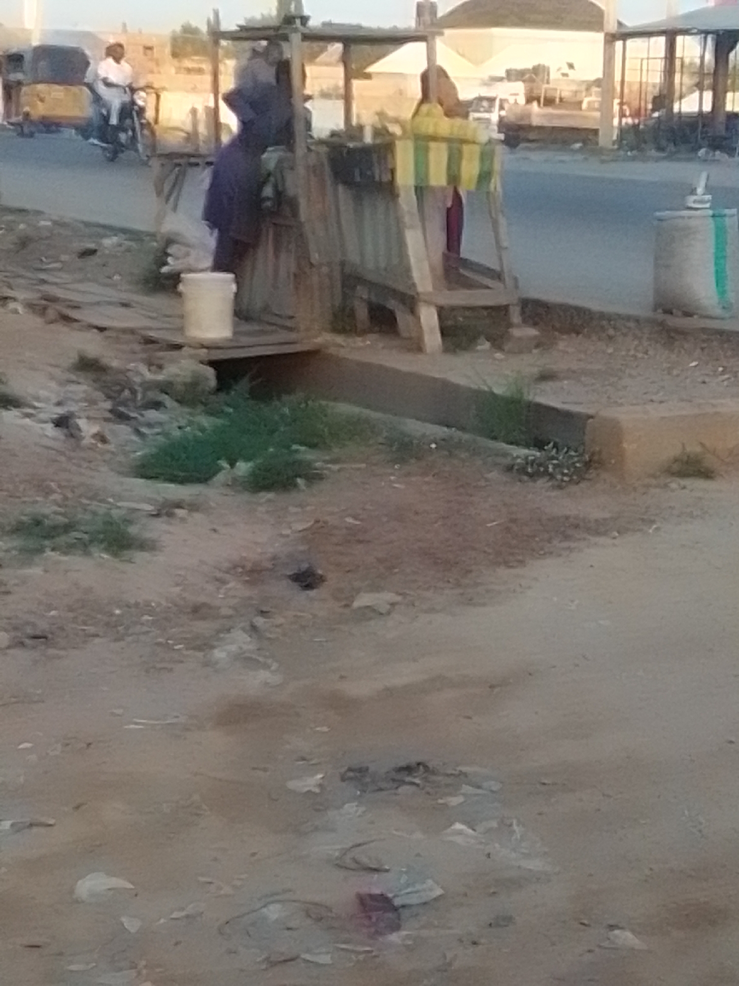 A street scene in Gombe, Nigeria, shows a person on a motorcycle with a yellow vehicle in the background. In the foreground, a wooden structure stands near a street. A white bucket is on the ground in front of the structure. A person wearing a blue garment and another person with a striped yellow and green cloth are visible near the structure. A large bag with a green stripe is on the right side of the image. The ground is unpaved and littered with debris. The sky is bright, suggesting daylight.
