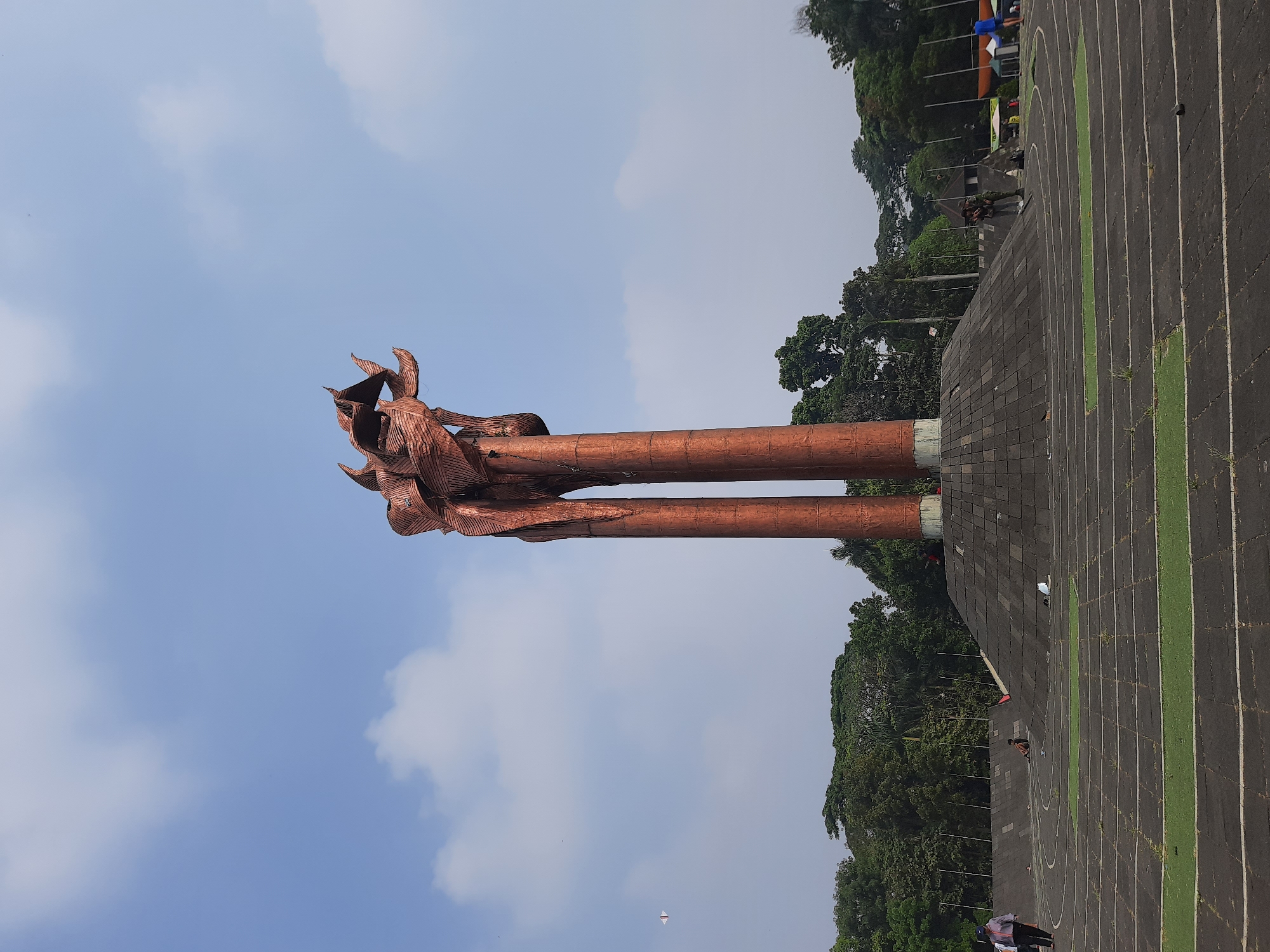 The image shows a wide shot of the Monumen Pemuda (Youth Monument) located in Bandung City, Indonesia. The monument is a tall, copper-colored sculpture with two main pillars supporting an abstract, multi-layered top. It stands on a raised, circular, tiered platform made of dark stone. The area surrounding the monument appears to be a park or open space with a running track visible in the foreground. The track is dark gray with white lines and some green sections. Several trees with lush green foliage form a backdrop behind the monument and the platform. The sky above is a pale blue with scattered white clouds. In the distance, a small white object, possibly a kite, is visible in the sky. There are a few people in the scene: a person in a blue shirt and white pants walks on the left side of the frame, another person sits on the ground near the monument, and a couple of individuals stand near the monument's base. The overall scene suggests a public outdoor space on a clear day.