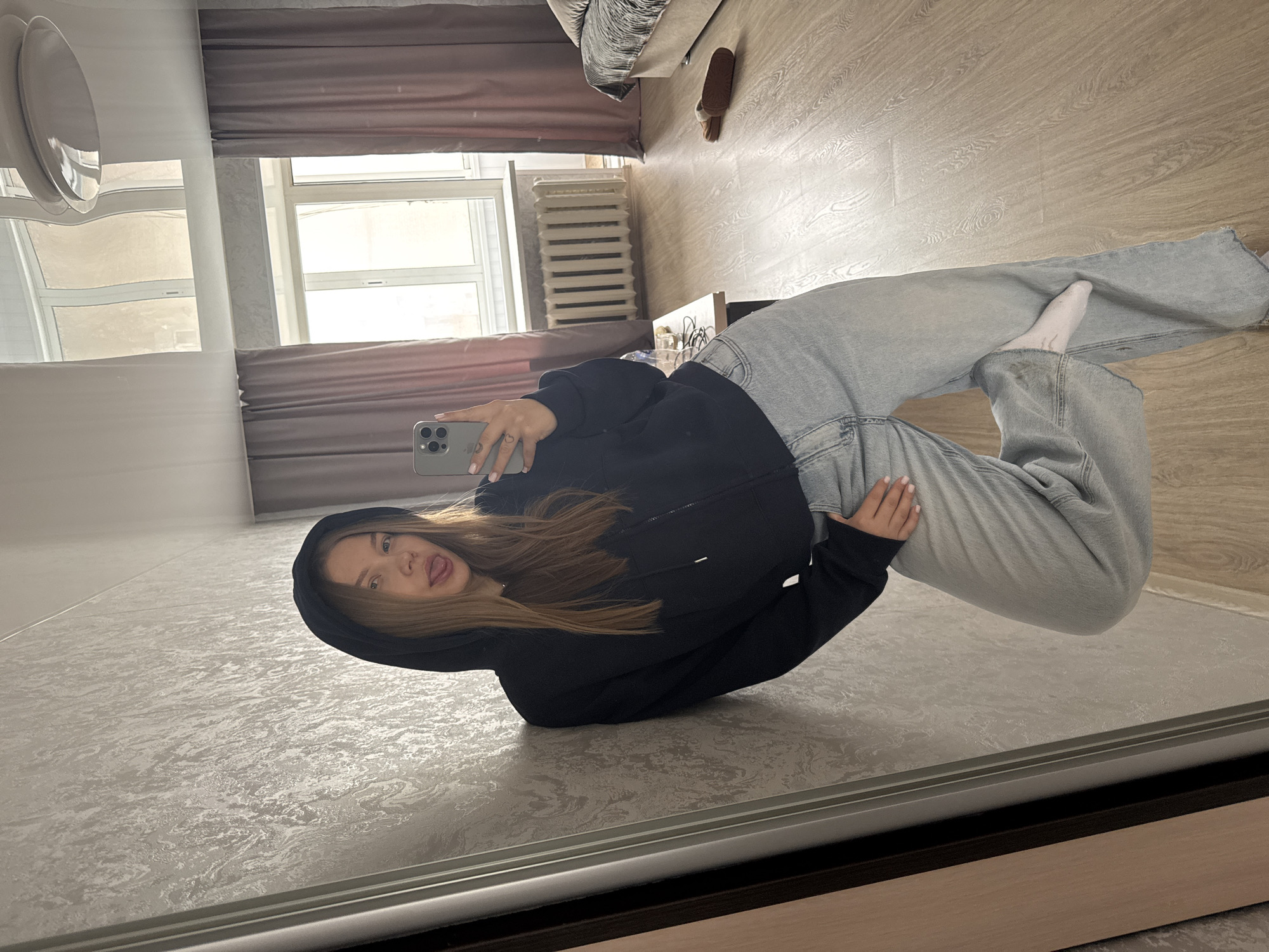 A woman is taking a selfie in a room. The woman is wearing a black hooded sweatshirt and light-wash jeans. She is sticking her tongue out. The room has beige walls and floors. A bed, window, and radiator are visible in the background. The city of Chaikovsky, Russia cannot be determined from this image.