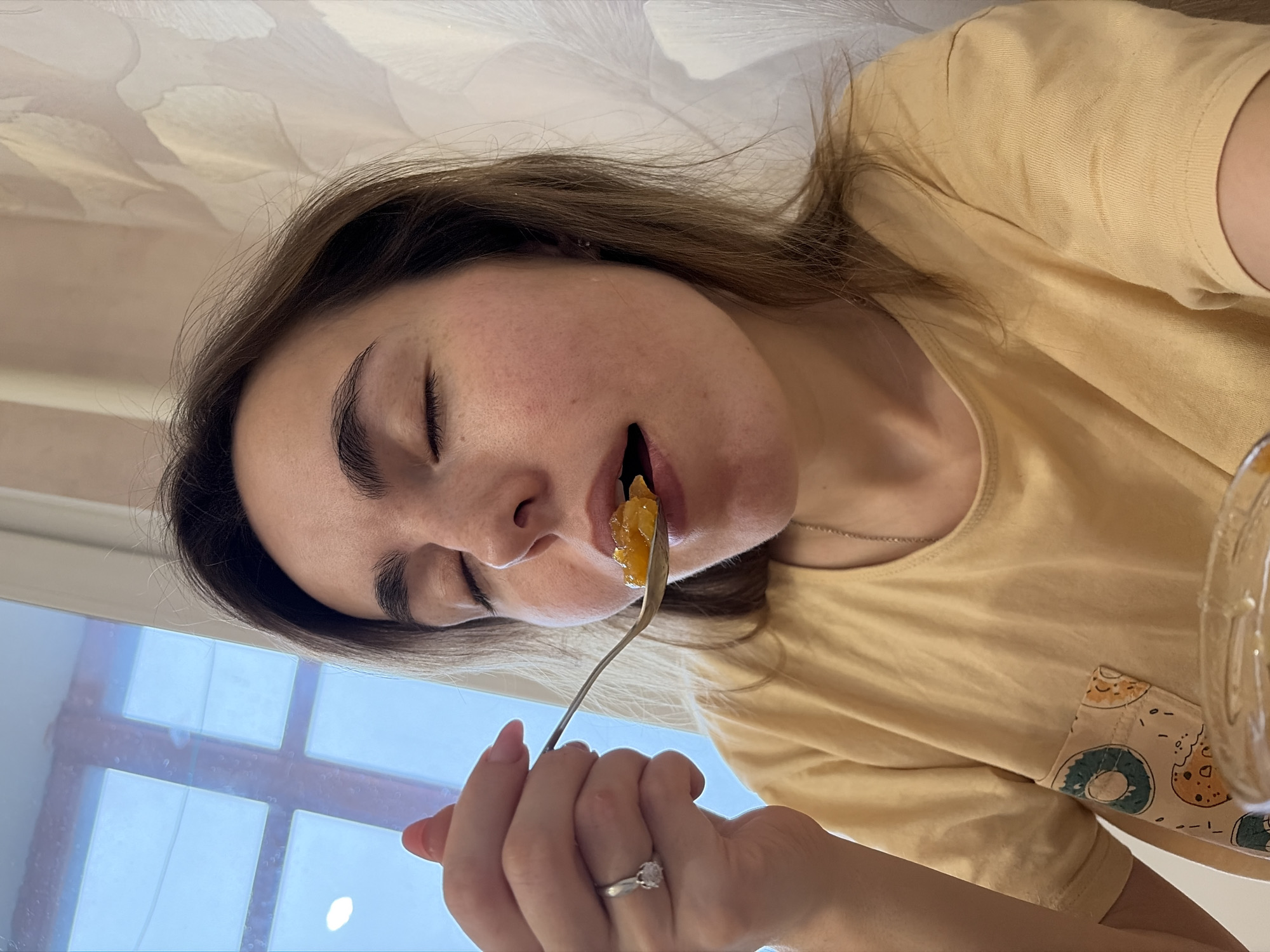 A woman is holding a fork with a piece of yellow food in her mouth. Her eyes are closed. She has brown hair. She is wearing a yellow shirt. She has a ring on her finger. A window is in the background. The location of the scene is not provided.