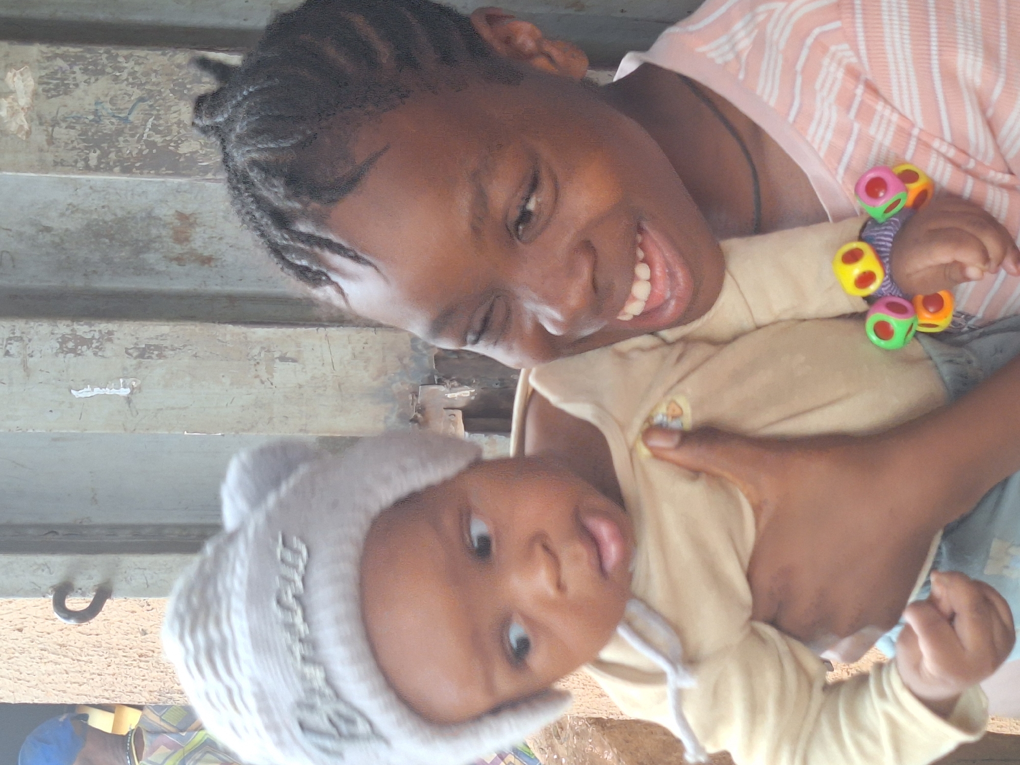 A smiling young woman with braided hair holds a baby in a gray knit hat. The woman wears a striped pink and white shirt and holds a colorful toy in one hand. The baby wears a tan shirt and is sticking out their tongue. The background shows a rough, weathered metal surface with a hook visible. The location is Sabon Tasha, Nigeria.