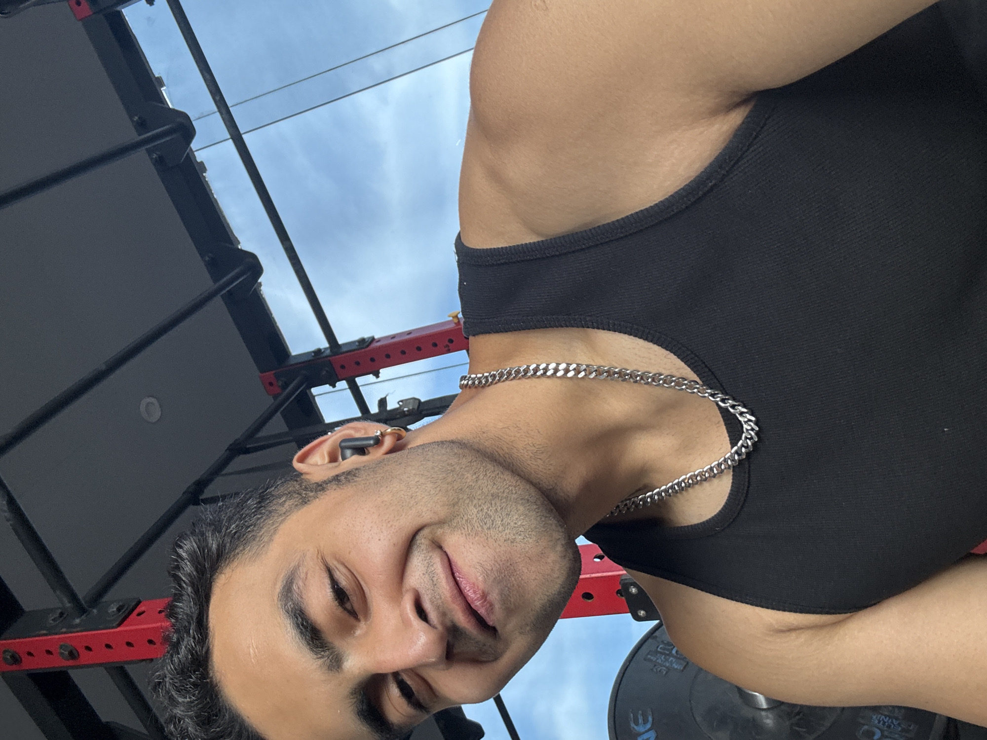 A man with short, dark hair and light brown skin is captured in a close-up selfie taken in a gym. He is wearing a black tank top and a silver chain. He has a small hoop earring in his left ear and is wearing a black wireless earbud in his right ear. He has a light beard and is smiling. In the background, there is a red and black weight rack and a portion of a weight plate. The sky is visible through a gap in the gym equipment. The image does not provide any information that would indicate the location is Kolkata, India.