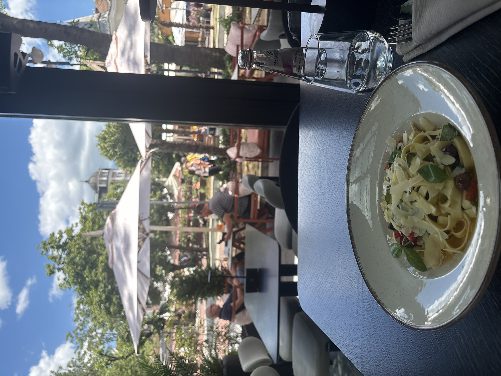 A close-up, eye-level, outdoor shot captures a plate of fettuccine pasta topped with shredded parmesan cheese, basil leaves, and what appear to be cherry tomatoes. The pasta is served on a light-colored ceramic plate with a textured rim. To the right of the plate are a clear glass of water, a bottle of San Benedetto natural still water, and a folded napkin with cutlery. The table is dark wood. In the background, the scene is blurred but suggests an outdoor dining area with several tables and chairs, some occupied by people. White umbrellas provide shade over the tables. Palm trees and lush green foliage are visible, along with buildings in the distance. The sky is blue with scattered white clouds. The overall impression is a pleasant, sunny day at an outdoor restaurant.