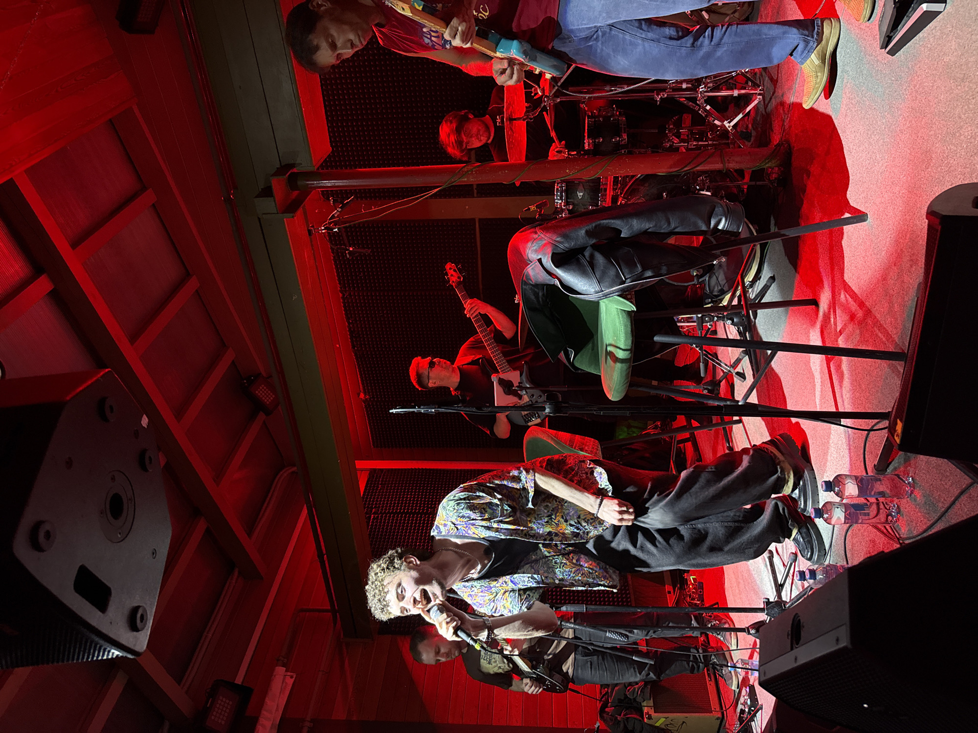 A vertically oriented image shows a band performing under red lighting. The singer is in the foreground, holding a microphone and wearing a patterned shirt with black pants and sneakers. Behind him, another band member plays a guitar. Further back, there is a bassist and a drummer visible. Two water bottles are on the floor next to the singer. A speaker is positioned on the left side of the image. The image was taken in what appears to be a music venue in Balashikha, Russia.