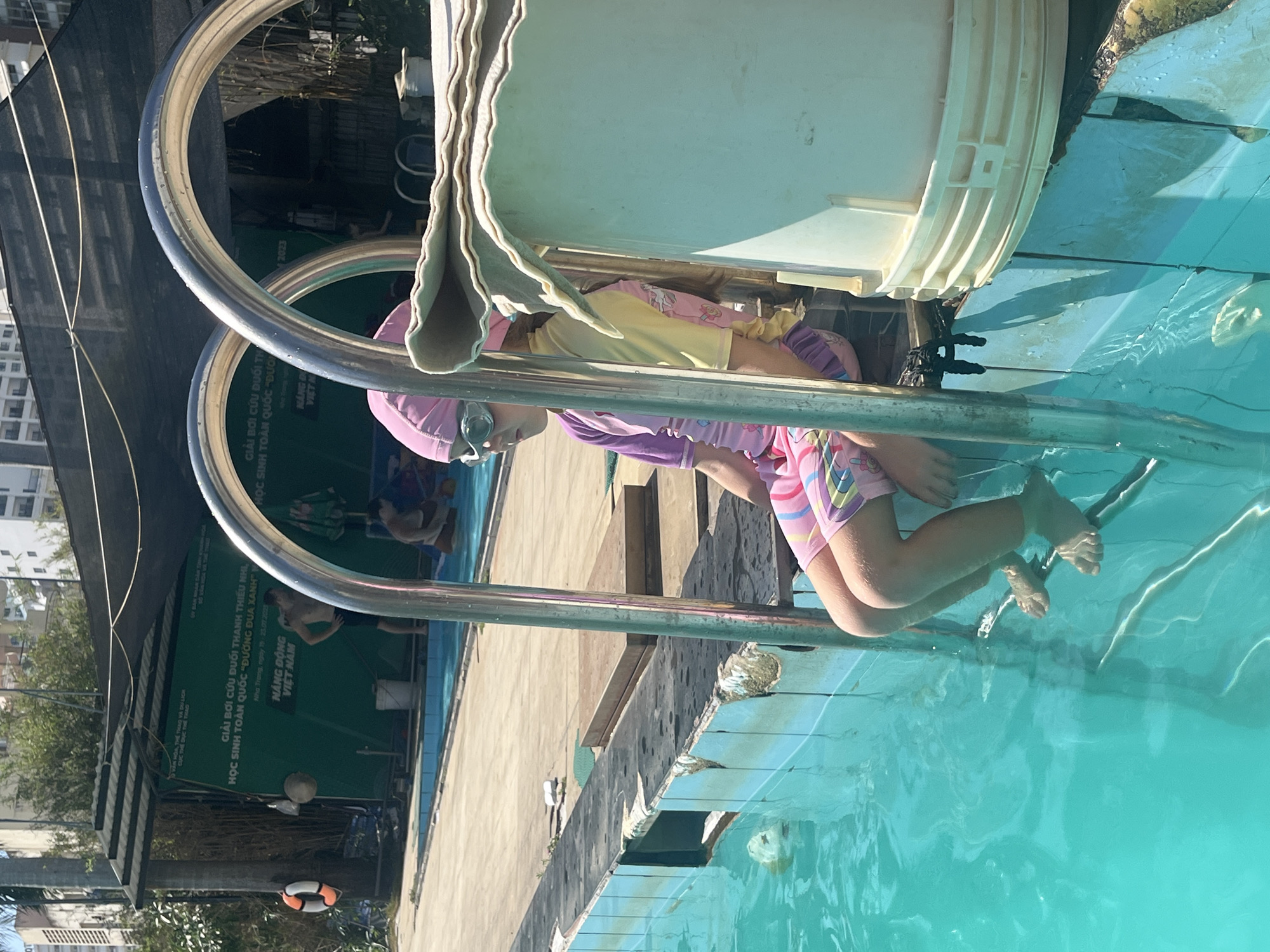A young girl wearing a pink swim cap, light-colored goggles, and a purple and pink patterned long-sleeved swimsuit sits on the second step of a metal pool ladder, with her bare feet submerged in clear turquoise water. Her gaze is directed to the left. To her immediate right, a light-colored plastic bucket with a beige cloth draped over its top edge rests on the pool deck. Behind her, a second child in a yellow swimsuit is partially visible.
The pool features worn light-blue tiled edges, exhibiting numerous chips, cracks, and visible grime. The surrounding deck is light grey concrete. In the background, a large green banner is displayed, obscured in parts by a black shade net. The banner's white Vietnamese text identifies a "National Youth Drowning Prevention Swimming Competition" ("GIẢI BƠI CỨU ĐUỐI THANH THIẾU NHI, HỌC SINH TOÀN QUỐC 'ĐƯỜNG ĐUA XANH'") held in "Nha Trang, ngày 19 - 23.09.2023" (Nha Trang, September 19-23, 2023), Vietnam. It also mentions "VĂN HOÁ, THỂ THAO VÀ DU LỊCH" (Culture, Sports and Tourism) and "CỤC THỂ DỤC THỂ THAO" (Sports Department), along with "NĂNG ĐỘNG VIỆT NAM" (Dynamic Vietnam).
To the left, a white lifebuoy with orange stripes hangs on a wooden post. Further back, a shirtless adult male stands near the pool edge. Other individuals are partially visible in the background, both in and out of the water. Multi-story buildings are visible in the distant background, beyond foliage, indicating an urban setting. The scene is illuminated by bright daylight, casting distinct shadows.