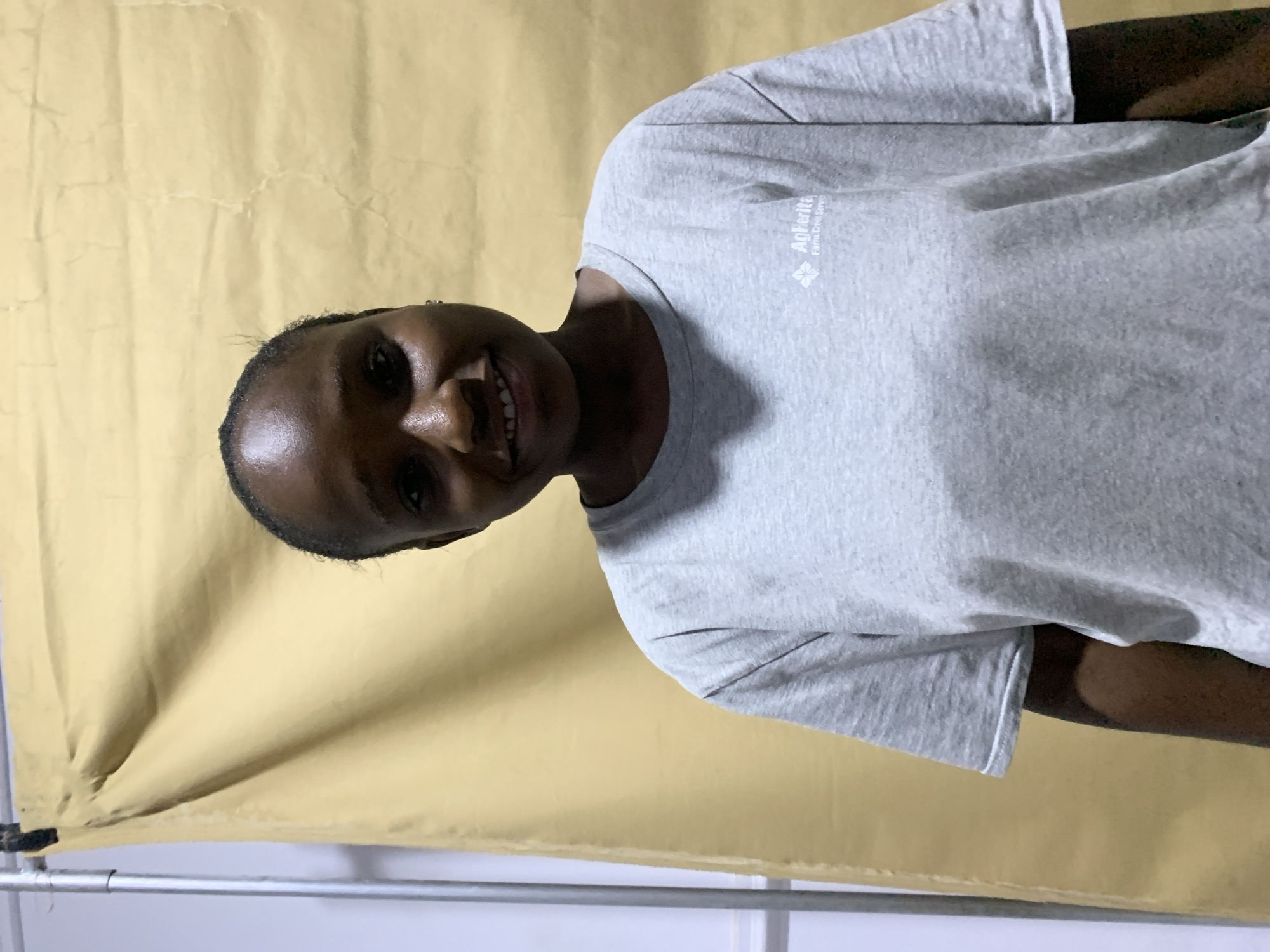 A medium shot shows a person with dark skin wearing a gray t-shirt, smiling. The person is positioned against a pale yellow backdrop. The location cannot be determined from this image. The city of Jos, Nigeria, cannot be confirmed based on the given information.