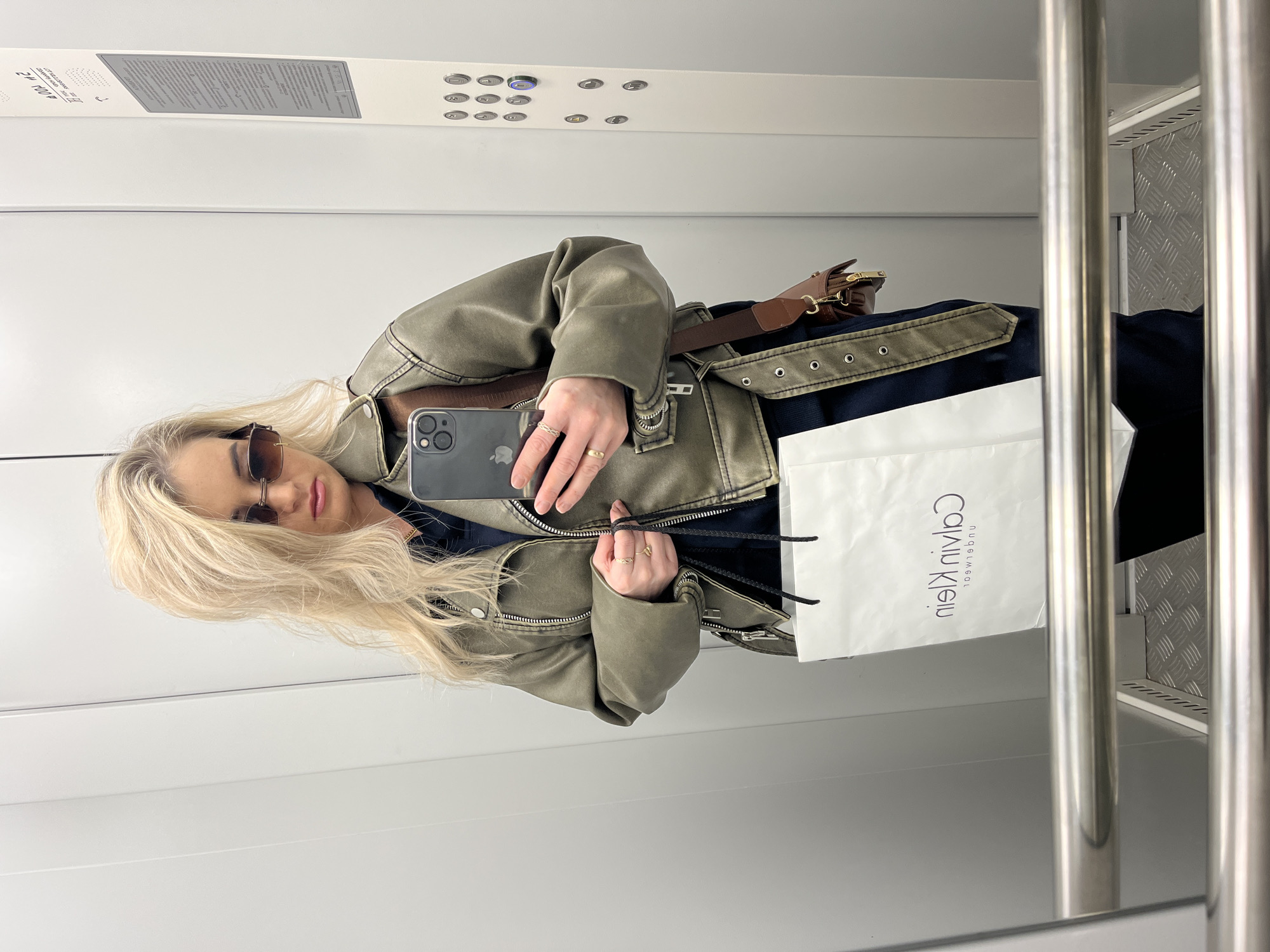 A person with long blond hair is taking a selfie in what appears to be an elevator. The person is wearing sunglasses, a gray-green leather jacket with a belt, and a dark blue garment underneath. A brown leather bag is strapped around the waist. The person is holding a black smartphone with their left hand. In their right hand, they hold the strings of a white shopping bag that reads "Calvin Klein" in reverse. The elevator has white walls, metal handrails, and a panel of elevator buttons. A panel with writing is visible above the buttons. The specific location in the city of Chaikovsky, Russia is not discernable from the image.
