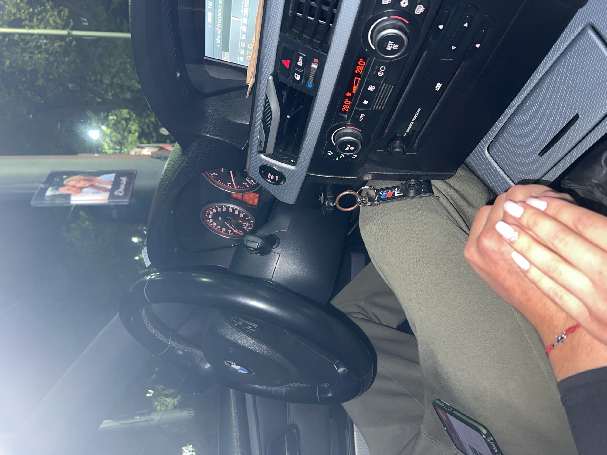 The image shows the interior of a BMW car at night, likely in Plovdiv, Bulgaria. The perspective is from the passenger seat, looking towards the driver's side and the front windshield.
A person's hands, with white manicured nails, are visible on the right side of the frame, resting on a khaki-colored leg. A red string bracelet with a silver cross charm is worn on one wrist. A smartphone is also resting on the leg. Through the windshield, blurry green foliage and artificial lights indicate an outdoor setting during the evening or night.
The car's dashboard is illuminated, showing an active infotainment system that displays "FM" radio frequencies from "88" to "102" MHz, along with the text "Manual frequency set" and "079 M." The climate control panel shows a temperature of "28.0°" and is set to "AUTO," with "MAX" and "REST" options visible. Other controls include a "START STOP" button, a hazard light button (partially visible, showing "OFF"), and air vents, one of which is marked "BMW." The gear indicator shows "D1," and the odometer reads "349459" km. The steering wheel, bearing a "BMW" logo and controls, is visible. Car keys with a "BMW M" keychain are also present. A small picture frame, featuring an image of two individuals and cursive text (partially OCRed as "Drama"), hangs from the rearview mirror. Additional visible text includes "NAVIGATION PROFESSIONAL" and "DVD" on the central unit.
