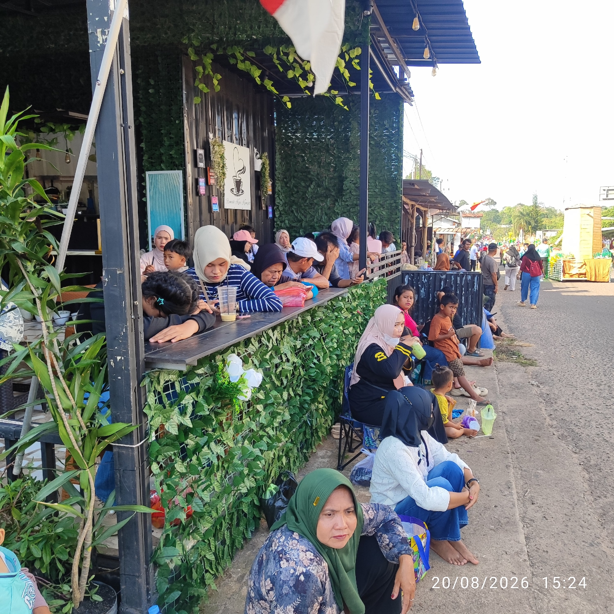 The image captures a street scene in Buding, Indonesia, likely in front of a cafe or food stall. The foreground features a group of people, primarily women and children, seated or leaning on a low barrier or ledge. Some are wearing headscarves, typical of the region. Several individuals are holding drinks. Behind them, a cafe-like structure with a dark corrugated metal exterior is decorated with artificial green foliage and some hanging lights. A red and white flag, possibly the Indonesian flag, is visible in the upper portion of the frame. The background shows a street with other stalls or buildings, trees, and more people walking. The lighting suggests it is daytime, possibly late afternoon, given the shadows. A date and time stamp "20/08/2026 15:24" is superimposed on the image.