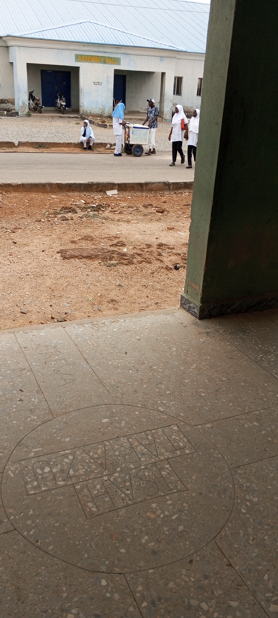 The image shows the exterior of a building labeled "ASSEMBLY HALL" in Keffi, Nigeria. A group of people are on the sidewalk and street in front of the building. One man is seated on the curb, while others are standing and walking, including a person in scrubs pushing a cooler on wheels. Other individuals are wearing hijabs or headscarves. The foreground shows a tiled or paved area with inscription "ISAIAH PARAKU ARENA." There's a street, some dirt/gravel ground, and a green pillar or wall on the right side of the frame. Two motorcycles are parked by the building. The sky is overcast.